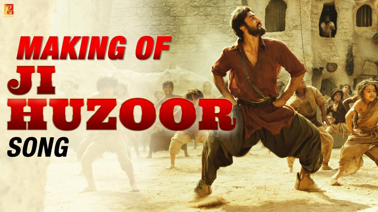 YRF 電影《Shamshera》「Ji Huzoor」歌曲場景 壁紙 — 一位留鬍鬚的男子身穿大地色長袍，在陽光明媚的沙漠村莊中充滿活力地跳舞，村民們從石頭建築中觀看。