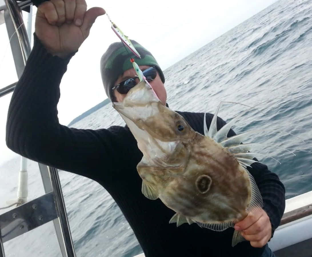  壁紙 — 一名漁夫在船上自豪地展示一條用彩色魚餌釣到嘅大魚，魚身佈滿尖刺，背景係波濤洶湧嘅大海。