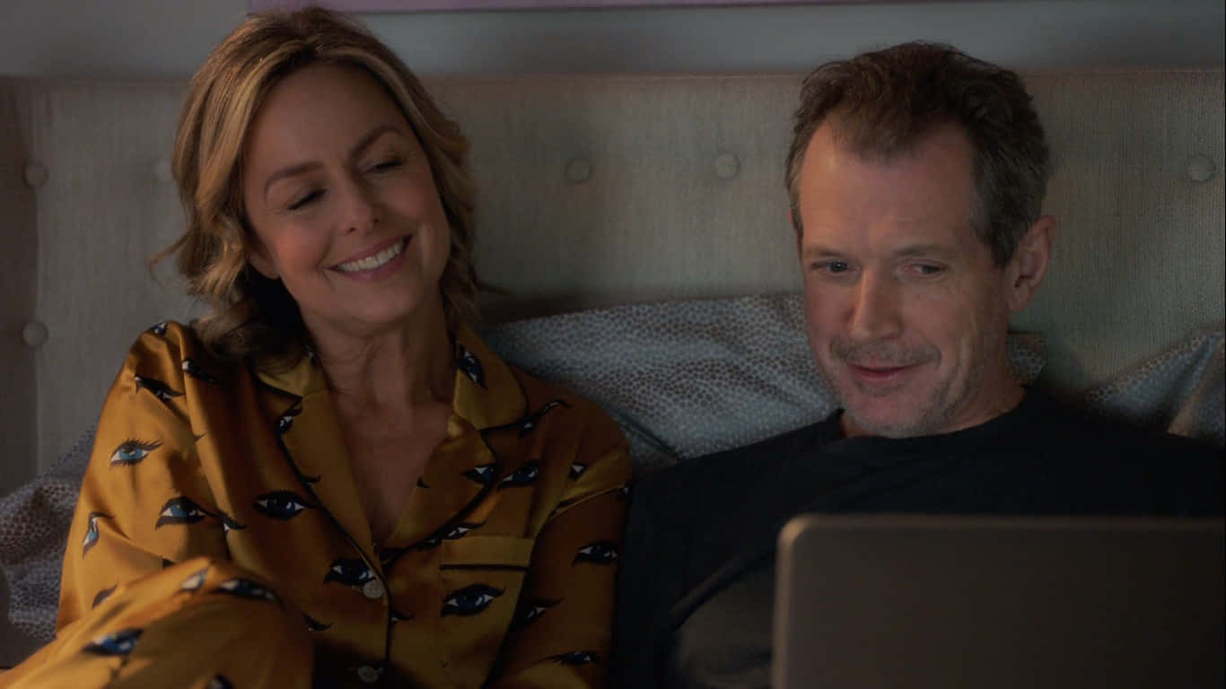 Melora Hardin 桌布 壁紙 — 一對微笑的情侶穿著睡衣在床上放鬆，一起在舒適的臥室裡看筆記本電腦上的東西。