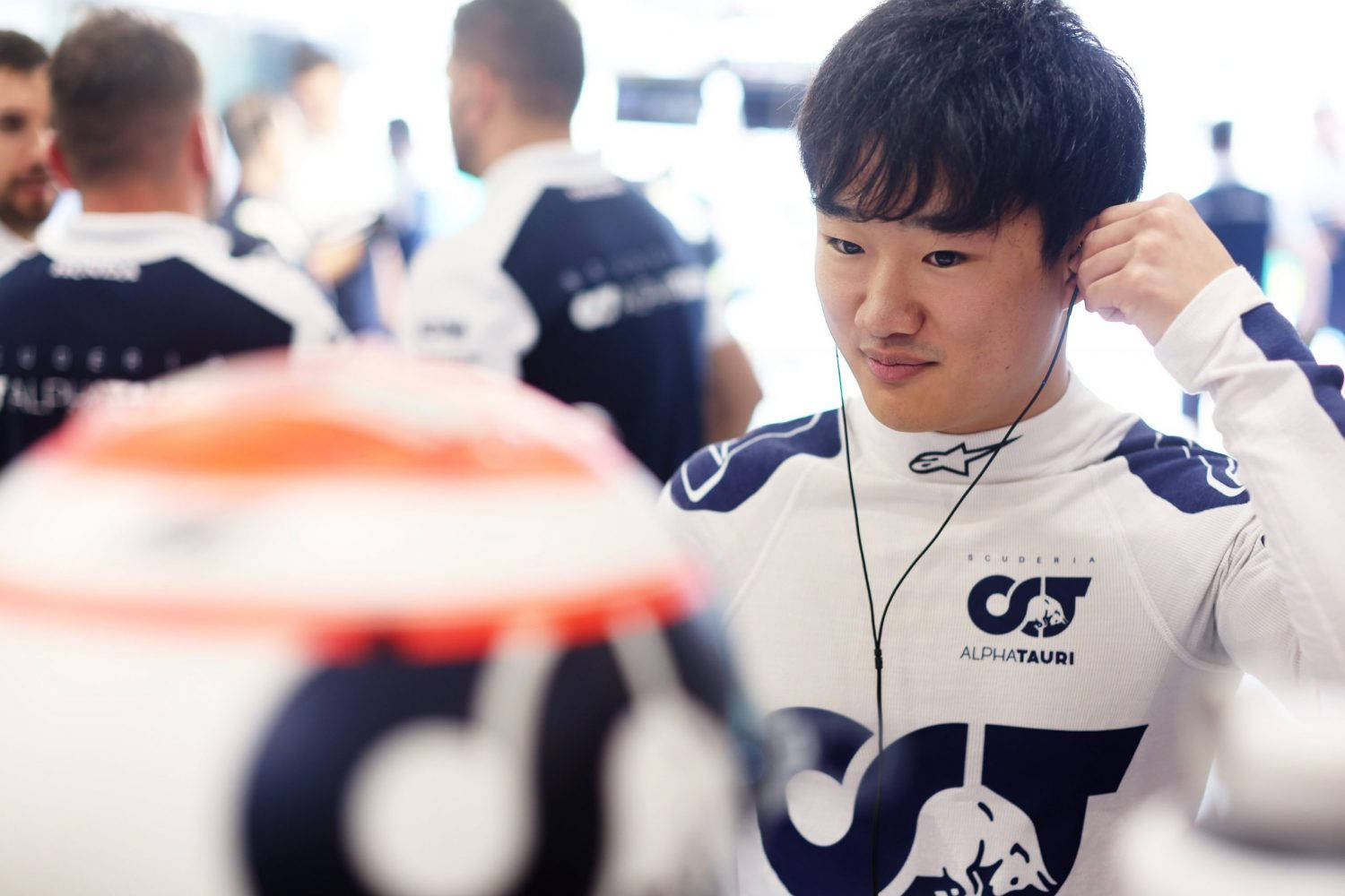 Yuki Tsunoda 賽前準備戴頭盔 壁紙 — 一位專注的F1車手身穿AlphaTauri車隊服裝，正在調整耳機，而賽車頭盔在前景模糊地放置。