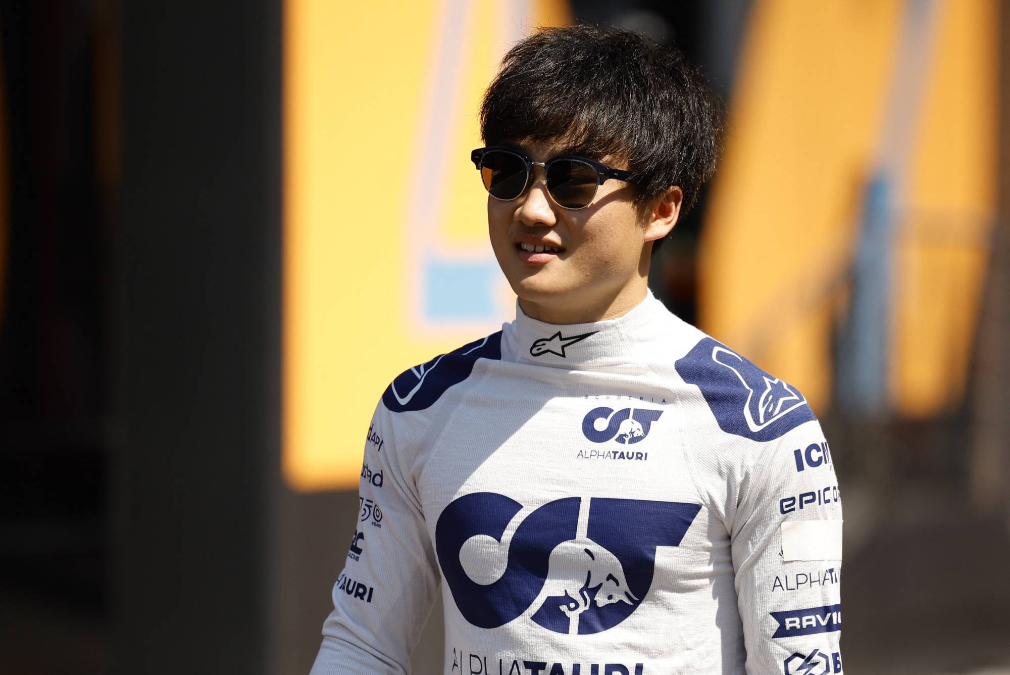 Yuki Tsunoda戴太陽眼鏡微笑 壁紙 — 一位面帶微笑的賽車手身穿AlphaTauri車隊賽車服和太陽眼鏡，自信地站在模糊的黃色背景前。