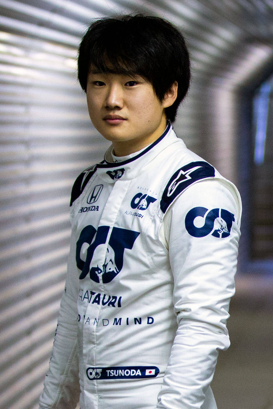 Yuki Tsunoda 在隧道內望向鏡頭 壁紙 — 一位年輕的亞洲賽車手身穿白色和藍色賽車服，自信地站在一個有波紋金屬牆的隧道中。
