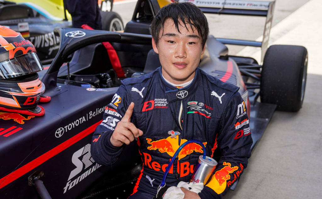 Yuki Tsunoda 旁邊的 Toyota Super Formula 賽車 壁紙 — 一位年輕的亞洲賽車手穿著Red Bull賽車服，在陽光明媚的賽道上坐在他的二級方程式賽車旁邊微笑。