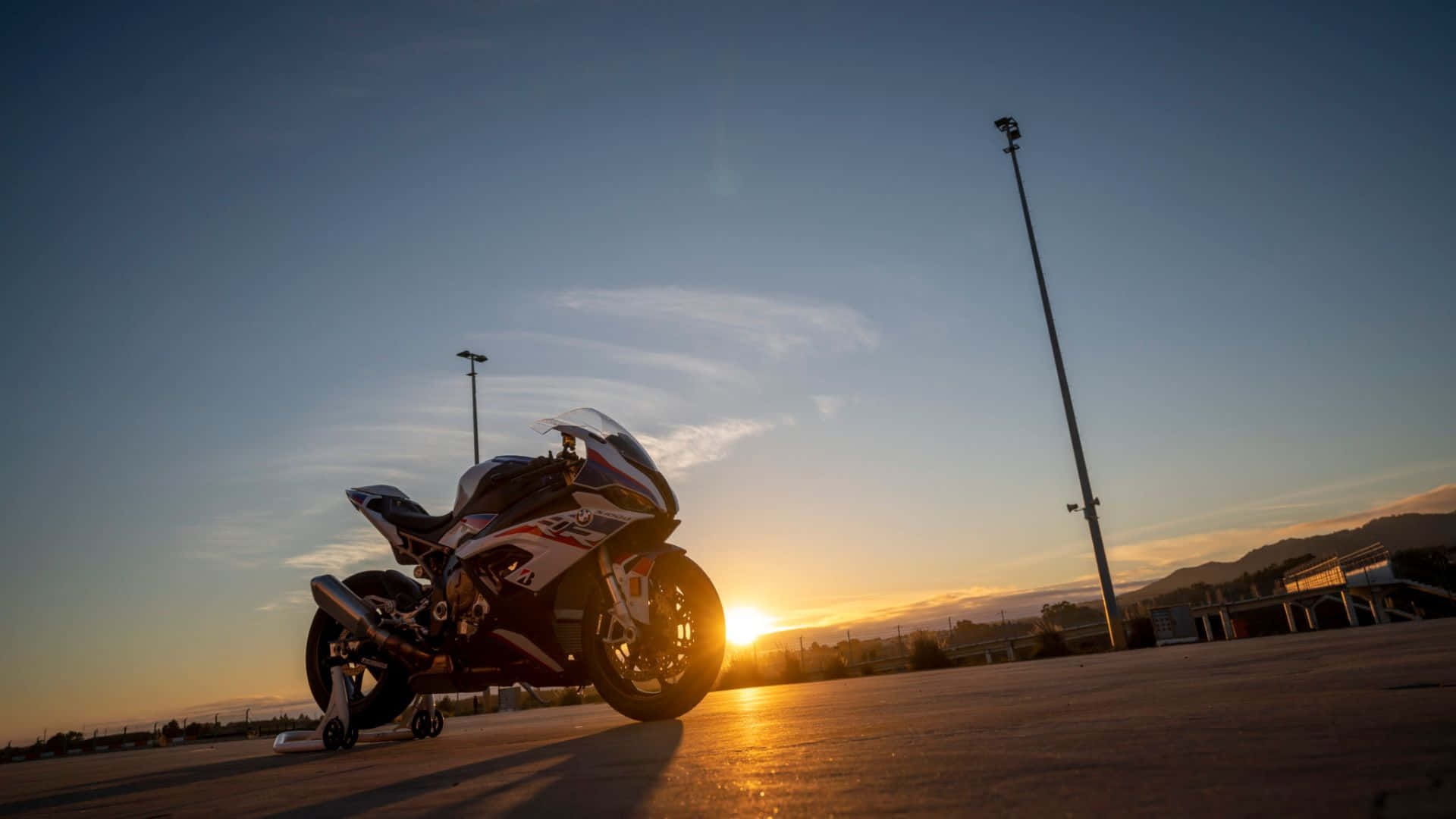  壁紙 — 一架運動電單車停泊在鋪砌的賽道上，夕陽在它背後落下，投下長長的影子與溫暖的金色光芒。