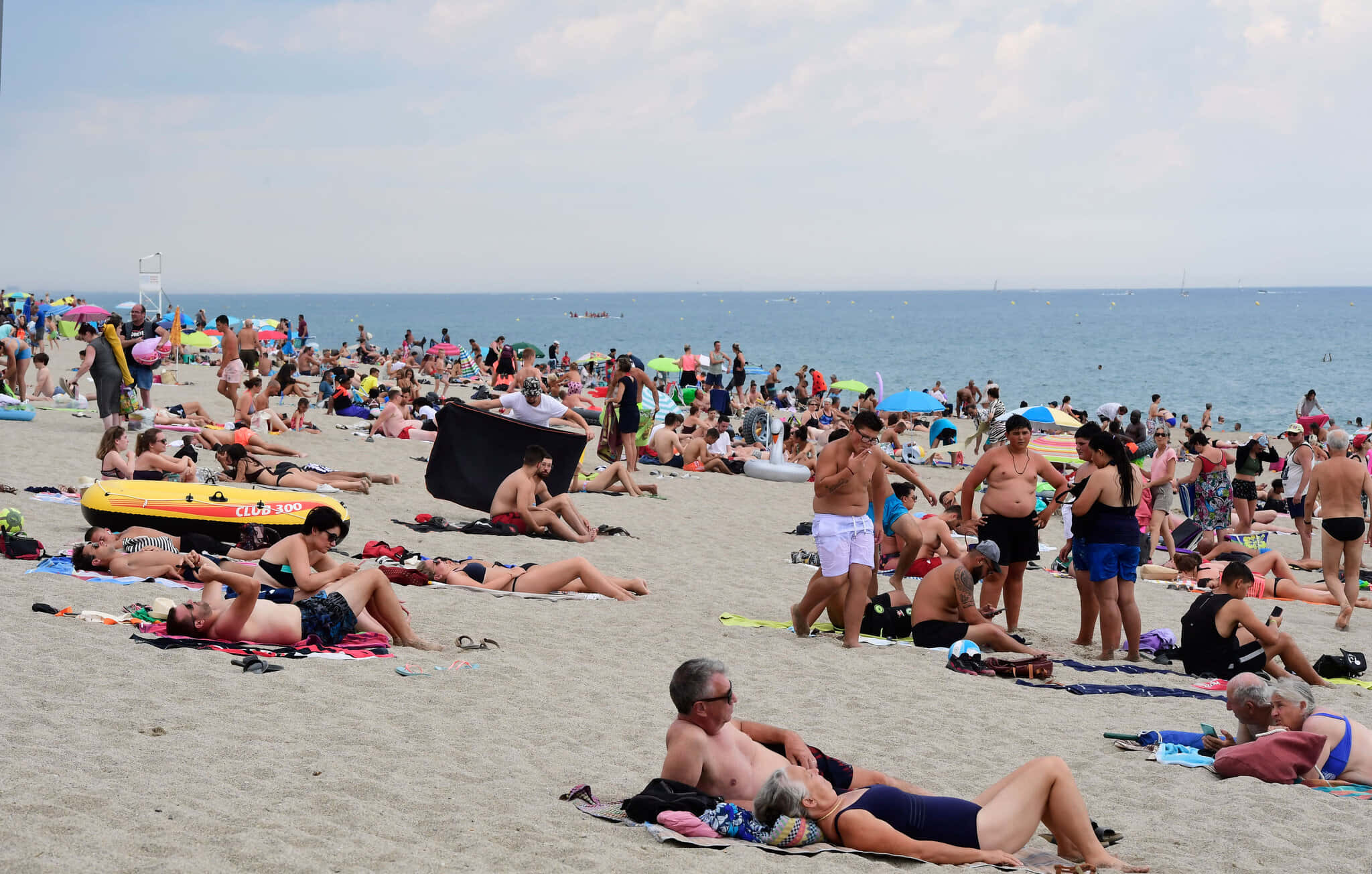 French Beach 桌布 壁紙 — 擁擠的海灘場景，陽光浴者在明亮的天空下於沙灘上放鬆，海洋延伸至地平線。