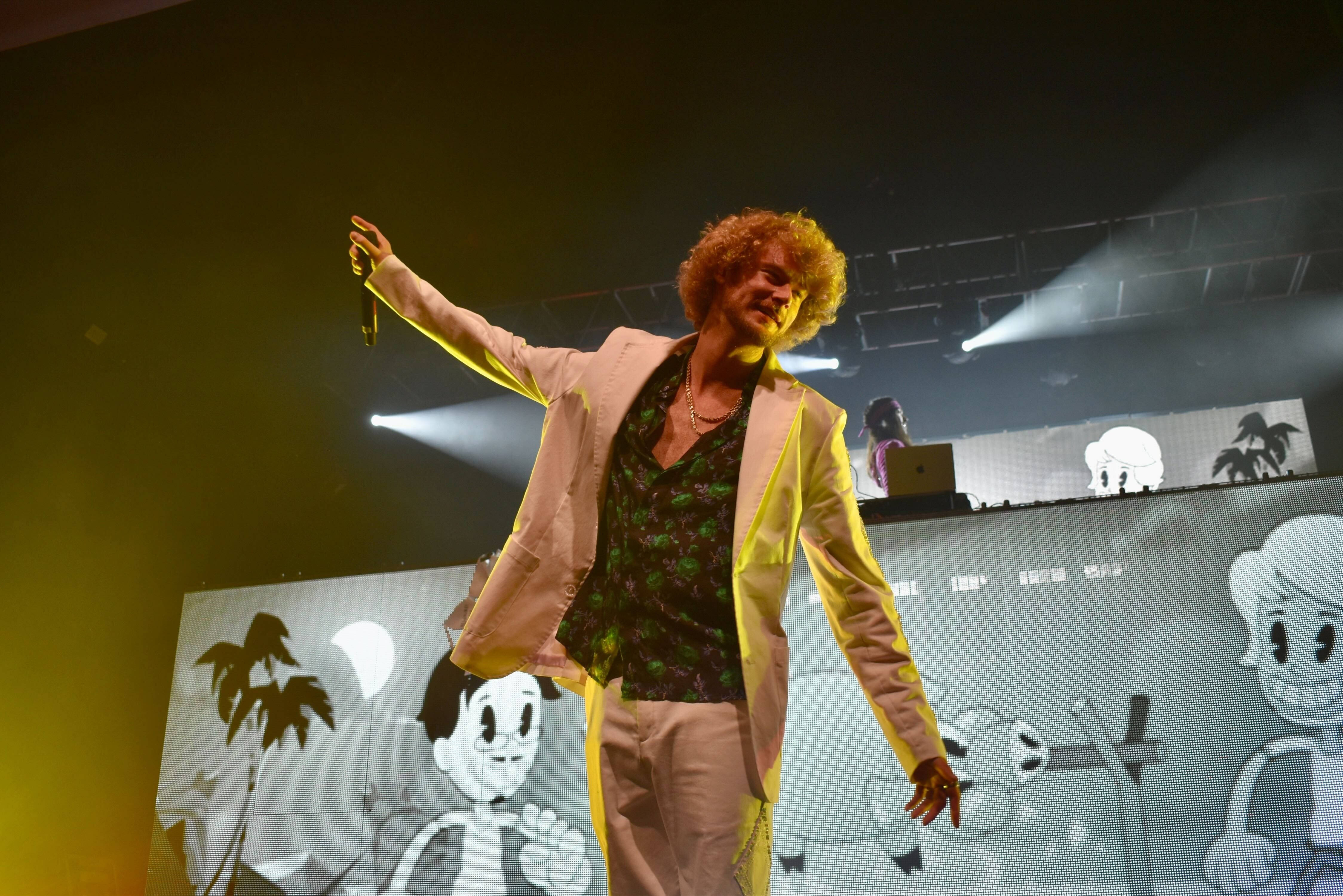 Yung Gravy 舞台表演 壁紙 — 一位捲髮表演者身穿粉紅色西裝，在明亮的聚光燈下於舞台上唱歌，背景有卡通人物。