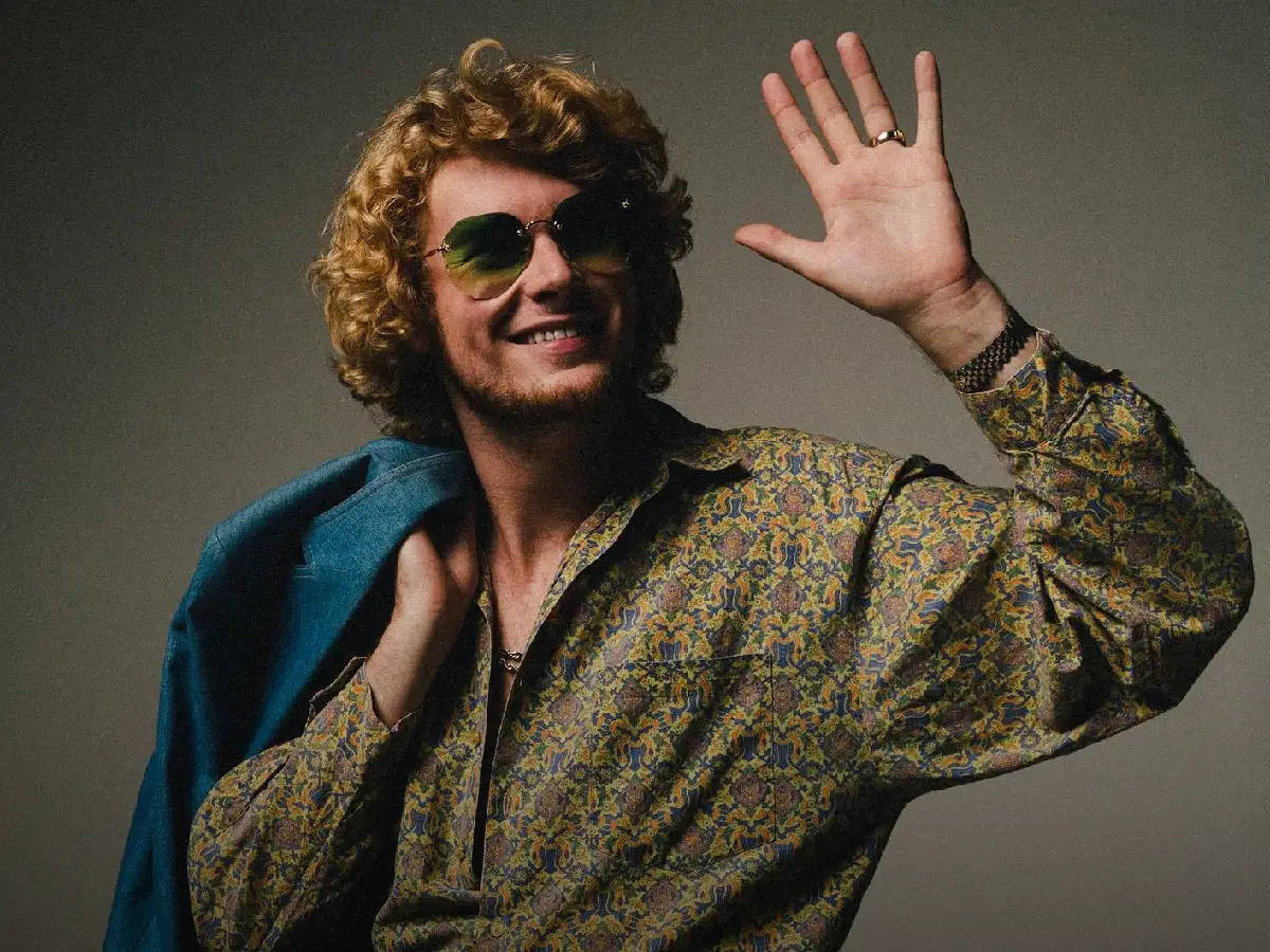 Yung Gravy 向粉絲揮手致意 壁紙 — 一個微笑的男人，捲髮戴太陽眼鏡，肩上掛著藍色外套，愉快地揮手。