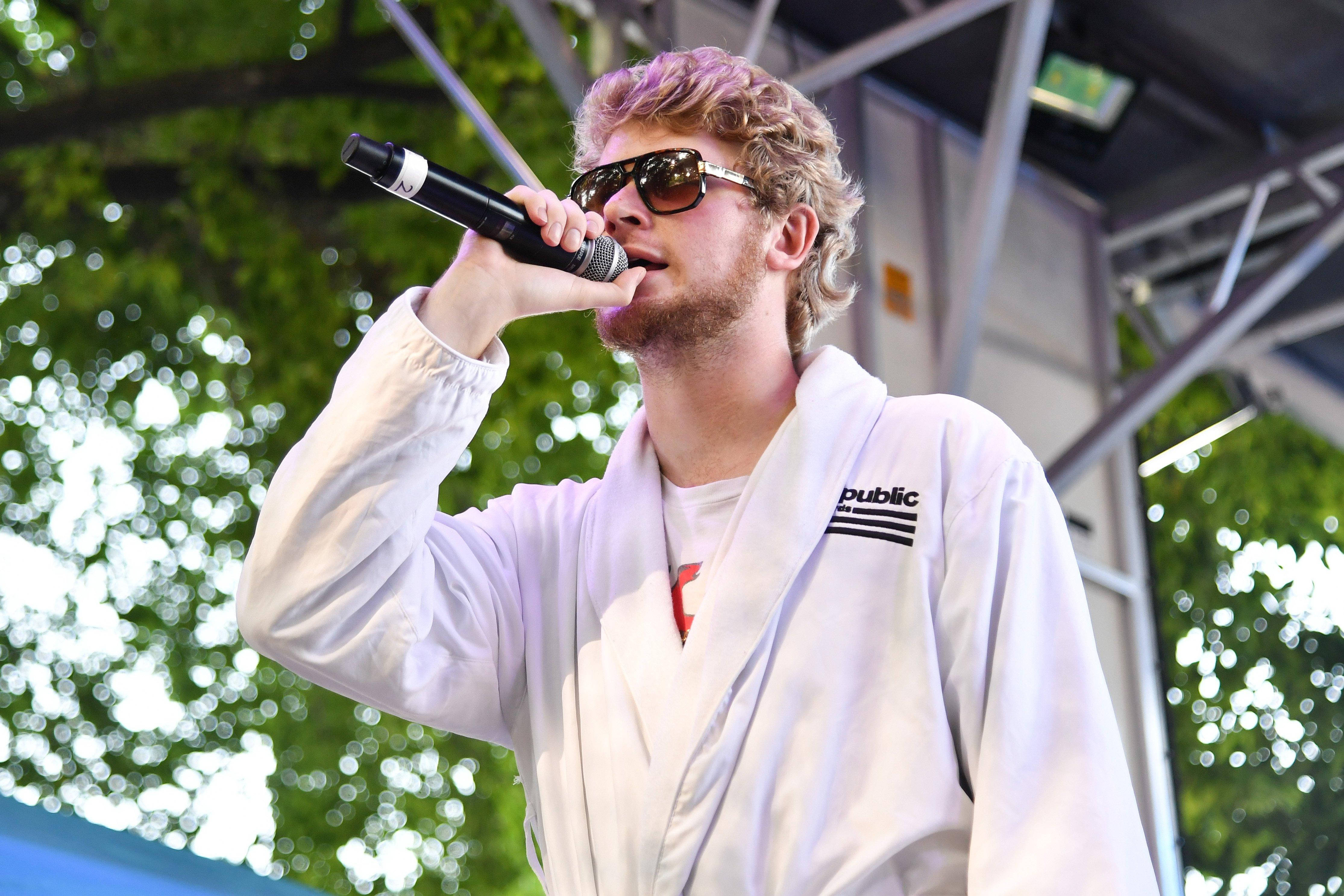 Yung Gravy 演出時刻 壁紙 — 一位金髮男子戴著太陽眼鏡和白色長袍，在身後有綠色樹葉的遮篷下對著麥克風唱歌。