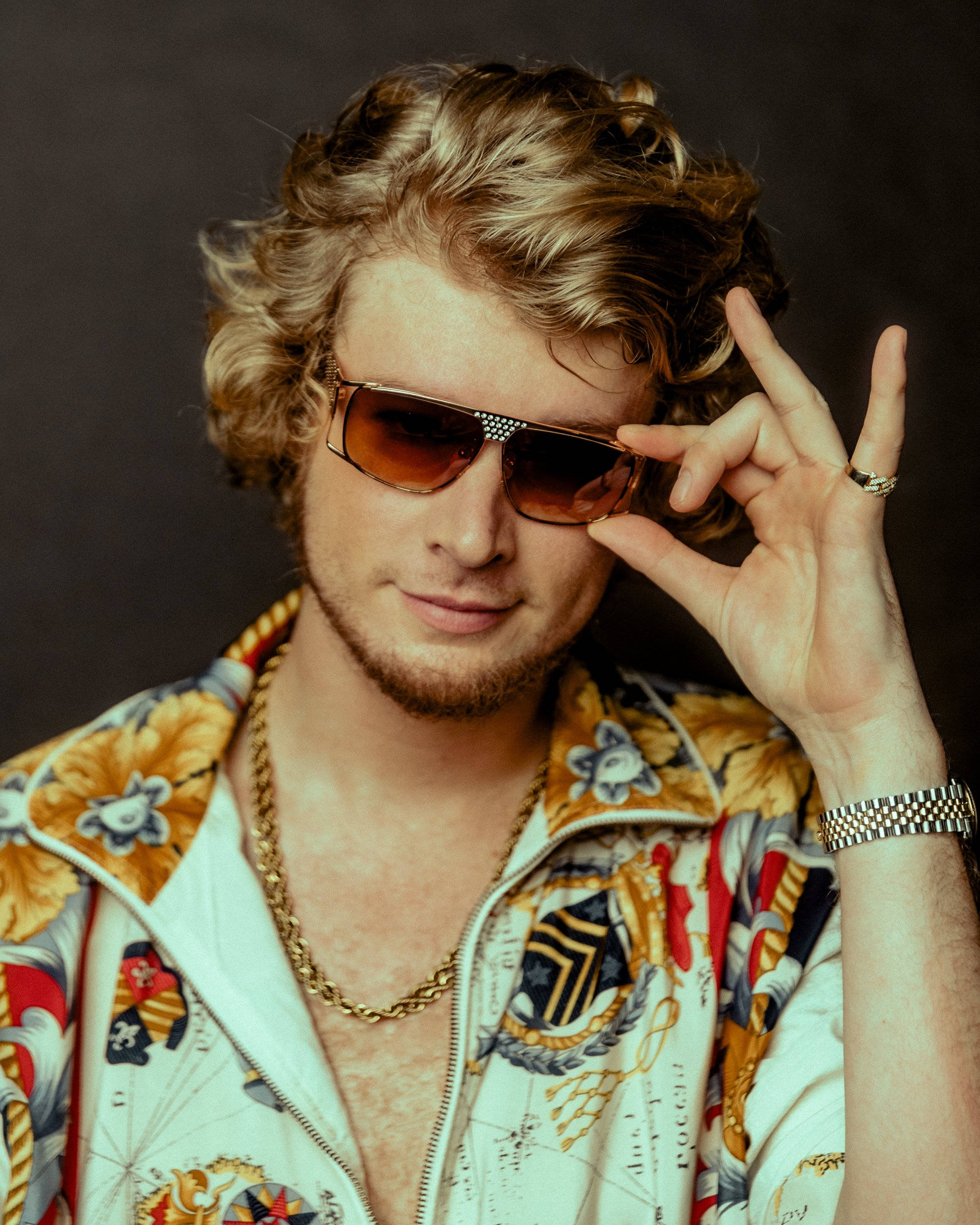 Yung Gravy 的潮流造型照 壁紙 — 一位時尚男士，金色捲髮，戴著華麗太陽眼鏡和圖案外套，比出和平手勢。
