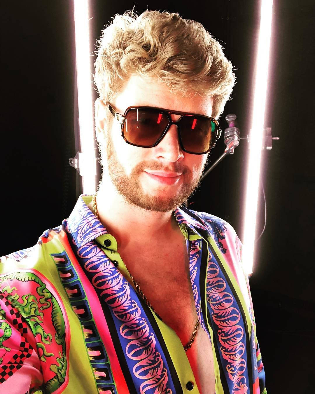 Yung Gravy 穿波浪髮型 壁紙 — 一個留著金色捲髮、戴著太陽眼鏡的男人，在黑色背景的明亮工作室燈光下穿著充滿活力的圖案襯衫。