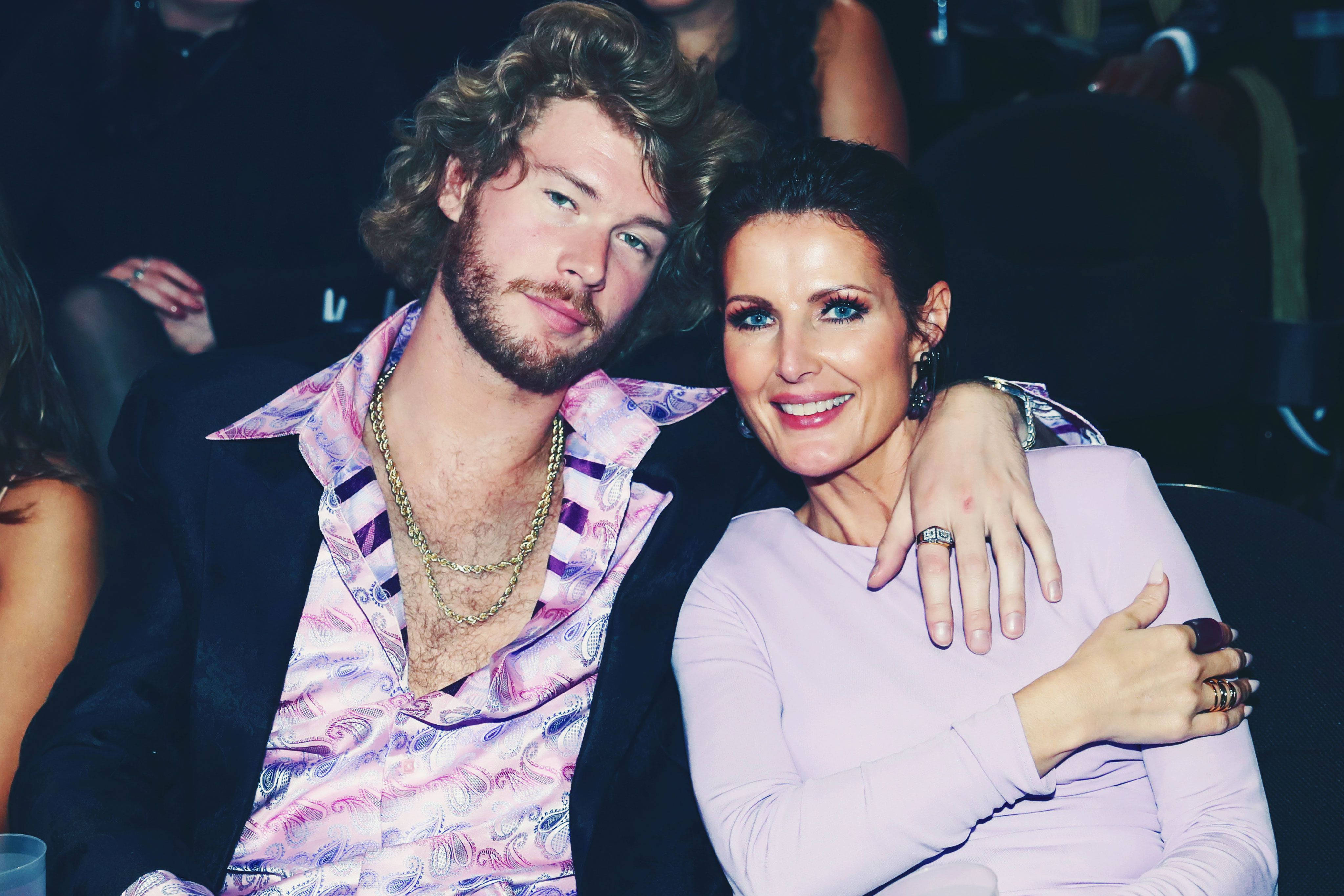 Yung Gravy 與 Sheri Easterling 壁紙 — 一位捲髮男士和一位穿著薰衣草色連衣裙的女士在燈光昏暗的活動中並坐，溫暖地微笑。