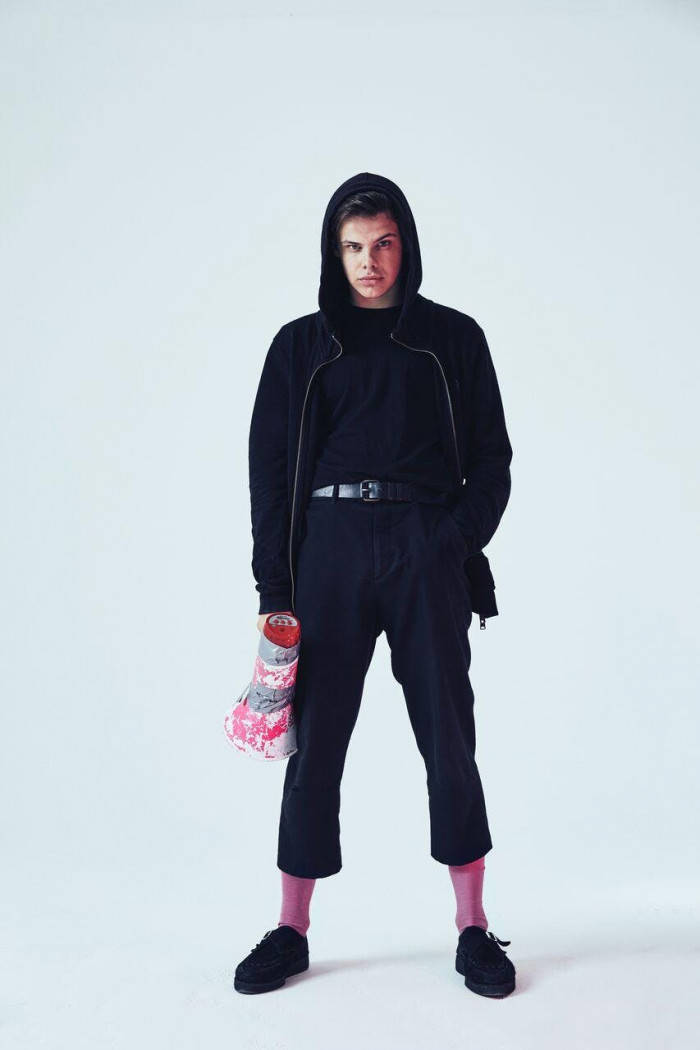 Yungblud 手持血跡麥克風手機壁紙 壁紙 — 一個穿著黑色連帽衫和褲子的人站在純白色背景前，拿著一個粉紅色和灰色圖案的袋子。