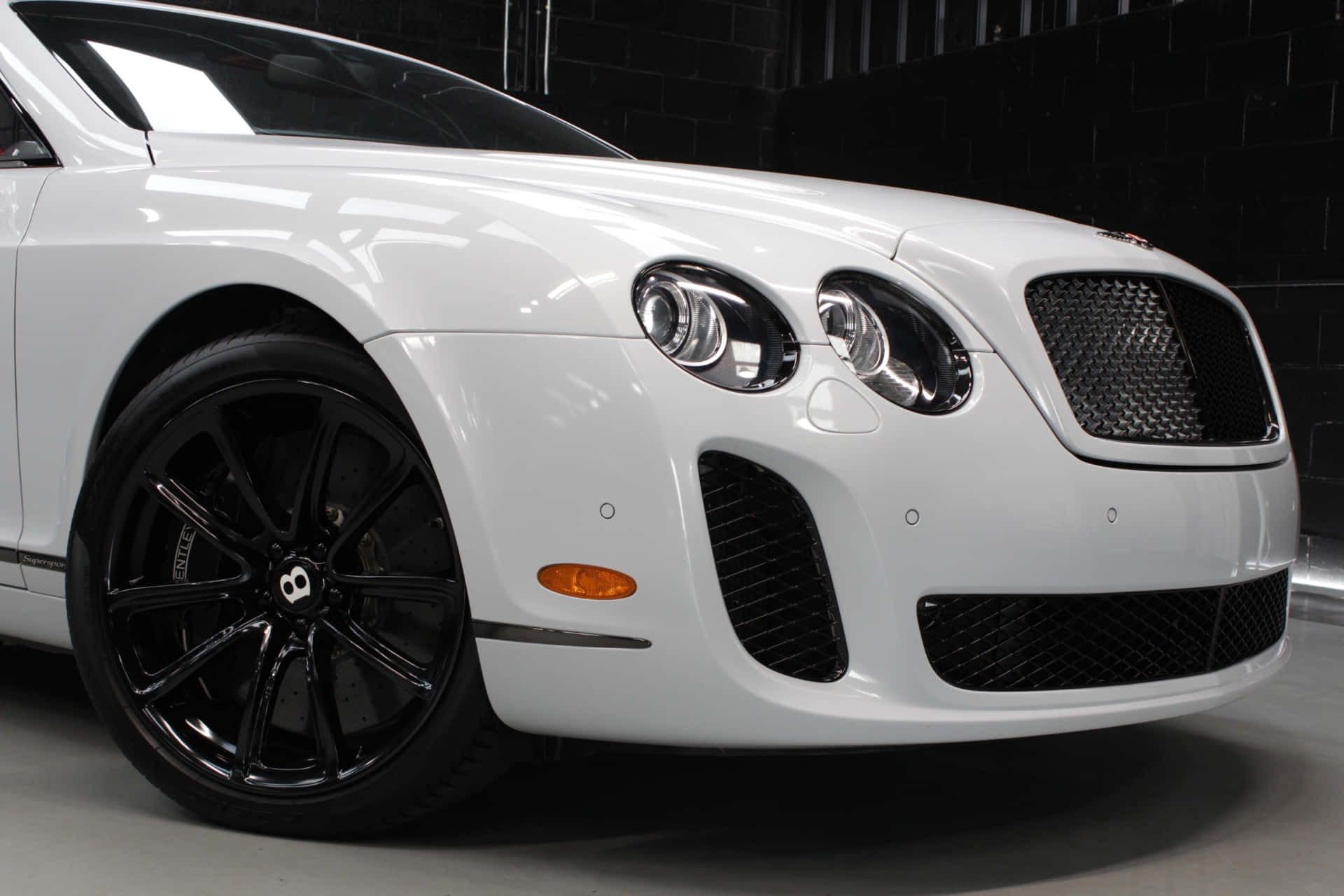 Bentley Continental Supersports 桌布 壁紙 — 一輛完美無瑕的白色 Bentley Continental GT 帶有黑色飾邊，在攝影棚燈光下對著深色磚牆閃閃發光。