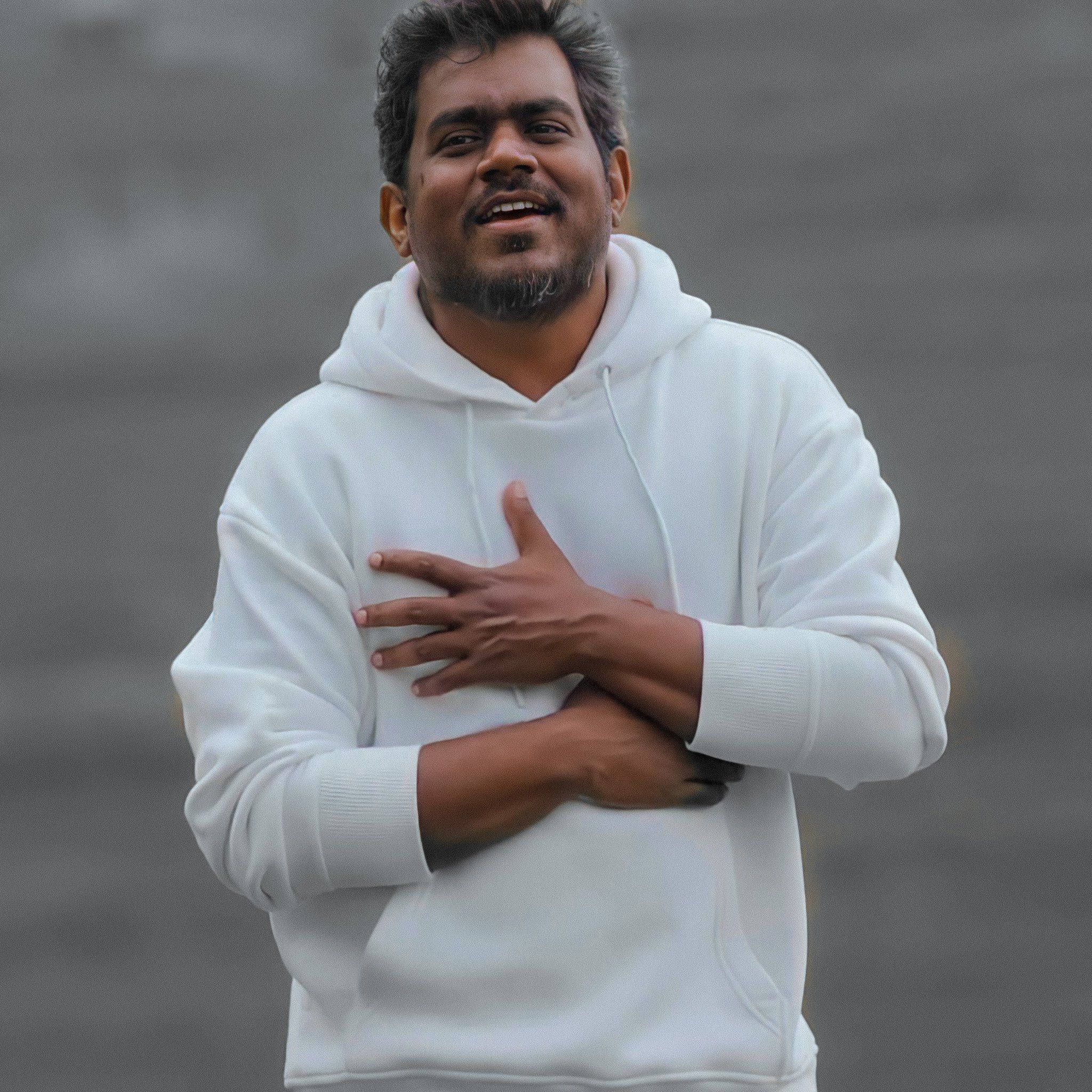 音樂大師Yuvan Shankar Raja穿時尚白連帽衫 壁紙 — 一名身穿白色連帽衫的男子，在模糊的灰色背景前，將手放在心口上，露出溫暖的笑容。
