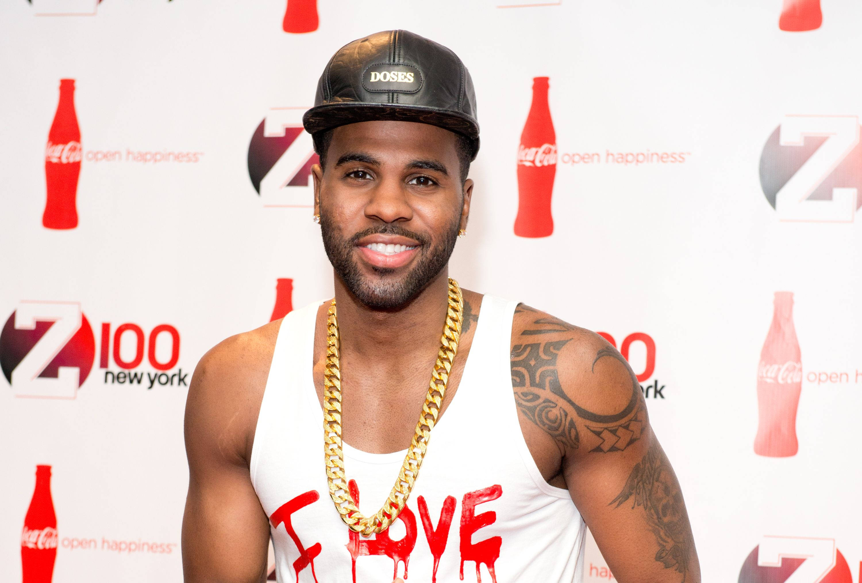 傑森·德魯羅出席 Z100 活動 壁紙 — 一名微笑的男子身穿白色背心及金鏈，在可口可樂品牌背景前擺姿勢。