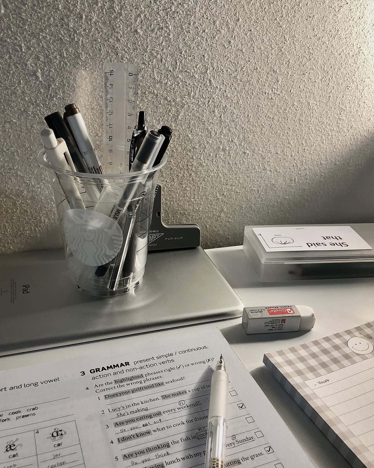  壁紙 — 一張雜亂的書桌，上面有一個透明杯裝著筆和尺，旁邊有語法練習紙和一本方格筆記本。