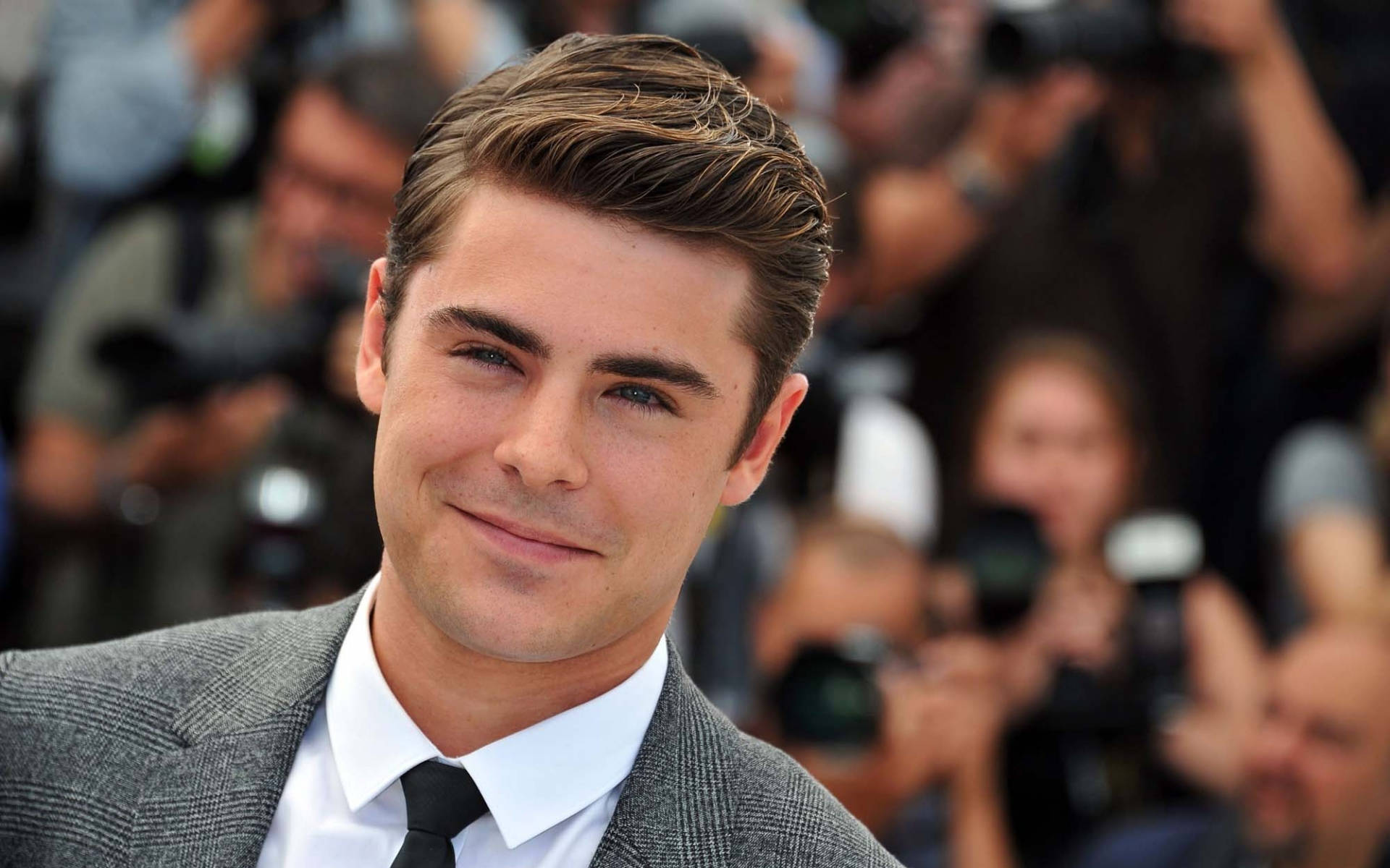 Zac Efron 穿著灰色西裝，在紅地毯活動中微笑，身後有攝影師。