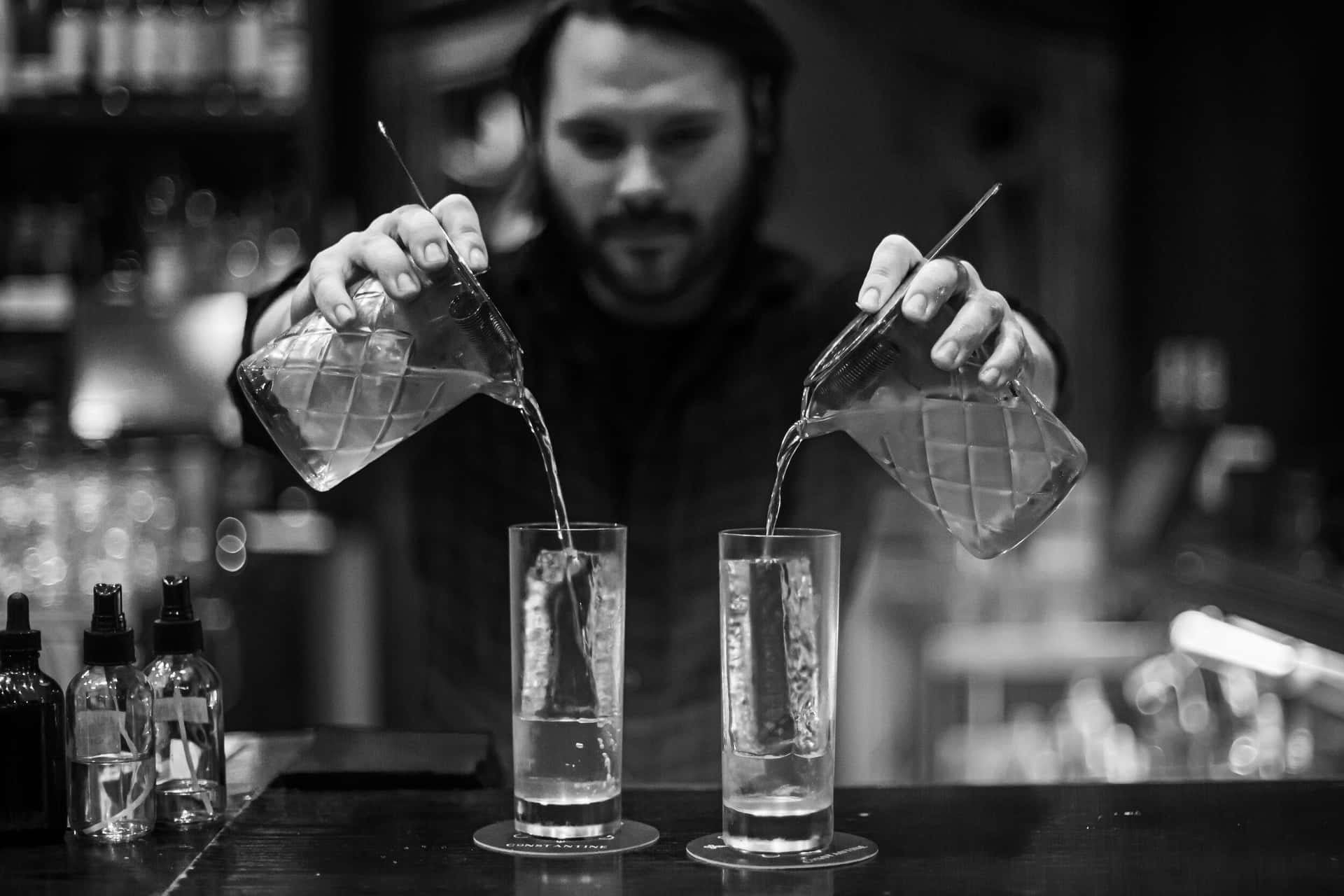  壁紙 — 在燈光昏暗的酒吧環境中，調酒師同時將兩杯飲料倒入裝有冰塊的高腳杯中。