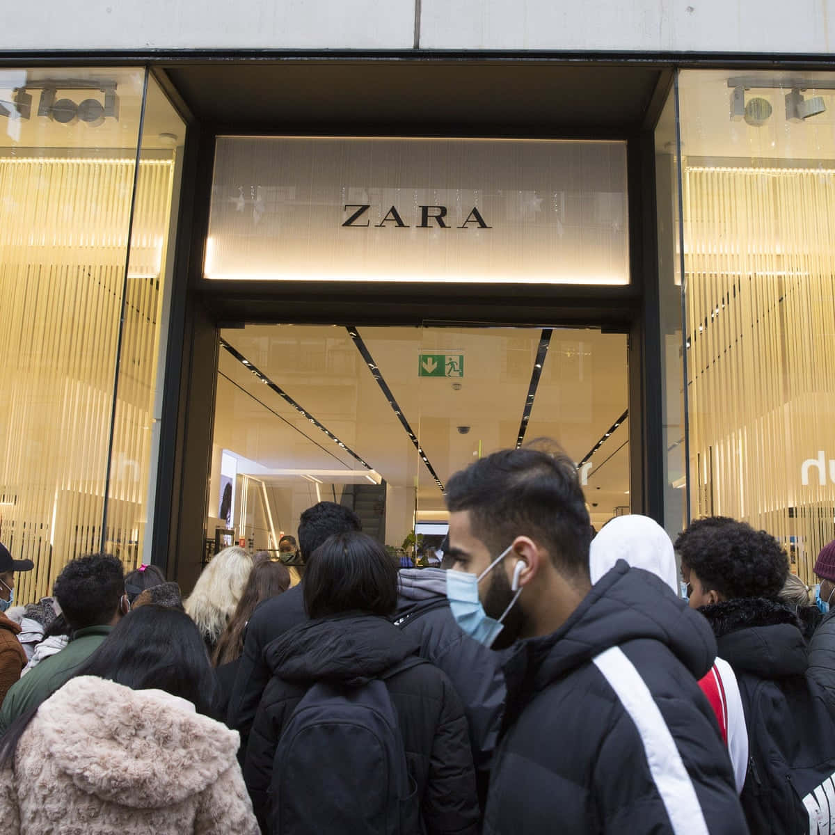 以 ZARA 最新系列刷新你的衣櫥 壁紙 — 戴著口罩的人群在明亮的 Zara 商店外排隊，商店擁有現代玻璃外牆和垂直燈板。