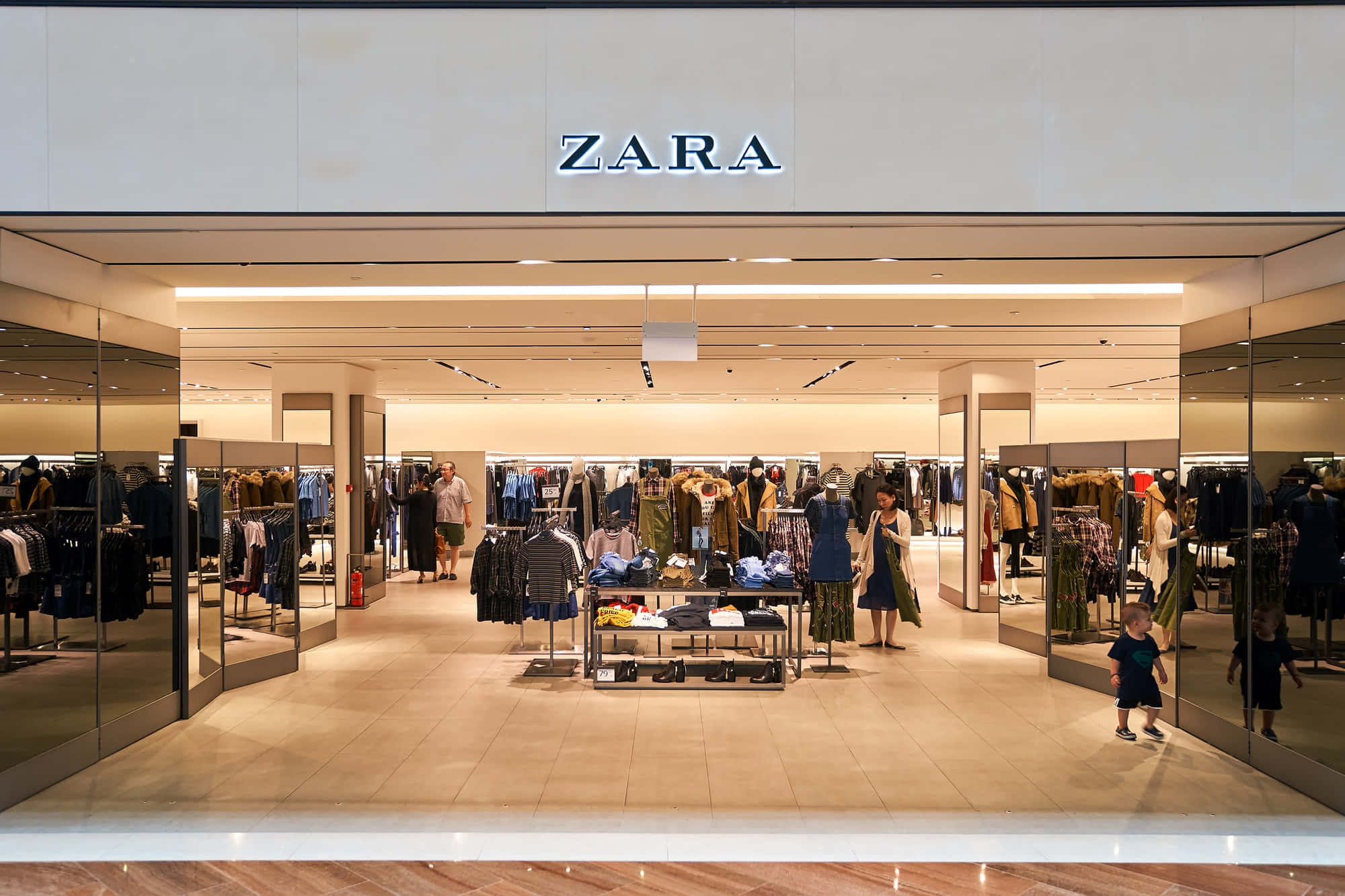 Zara 2021夏季最新時尚系列 壁紙 — 光線明亮的Zara店舖入口，玻璃牆反射出店內瀏覽衣架的購物者。