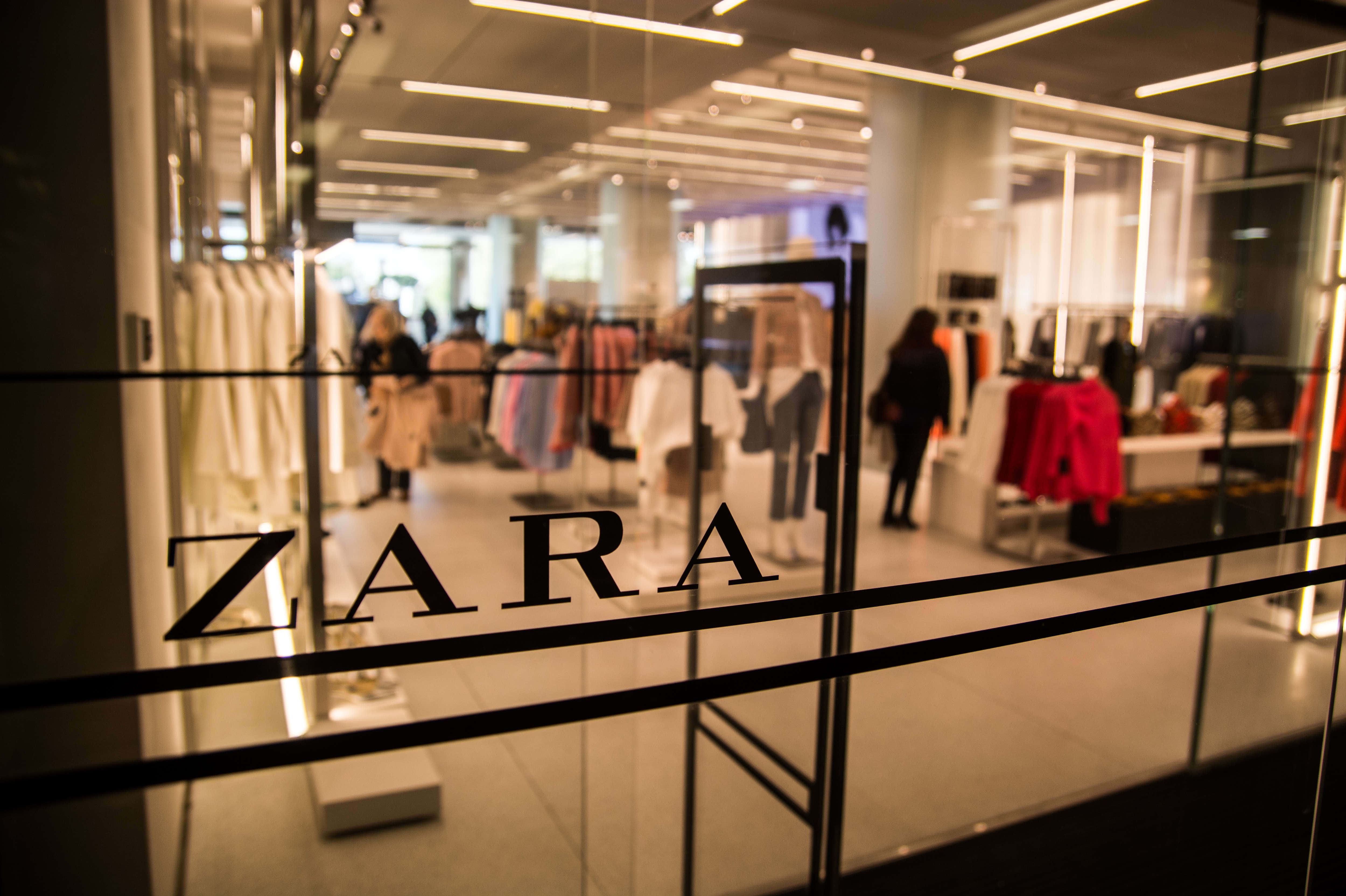 以Zara最新春裝刷新衣櫥 壁紙 — 透過Zara商店玻璃門的模糊視野，顯示顧客在明亮天花燈光下瀏覽色彩繽紛的服裝架。