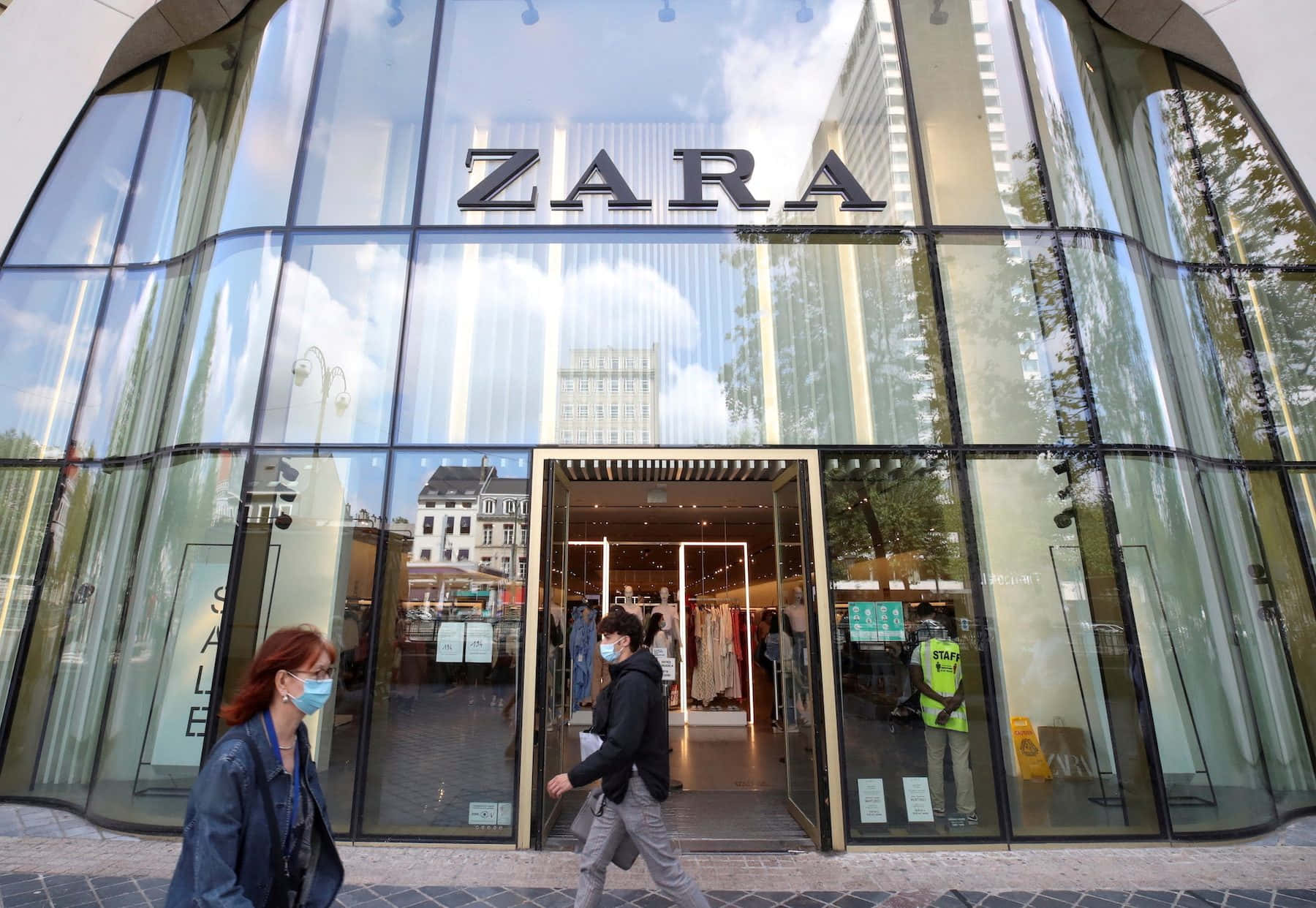 現代時尚與百搭造型 — Zara 壁紙 — 戴著口罩的人們走過一間現代化的玻璃幕牆Zara商店，倒映著明亮的天空和城市建築。