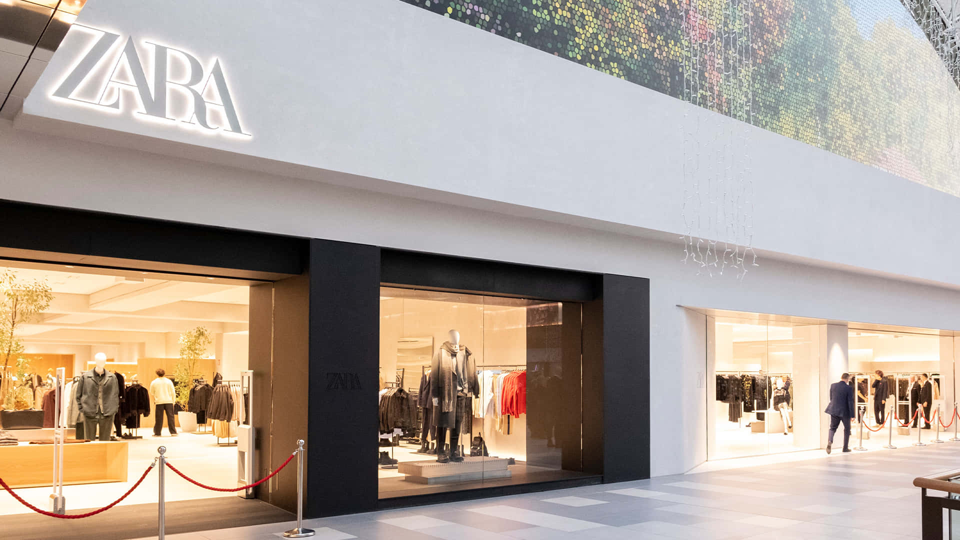 以Zara最新服裝提升秋季造型 壁紙 — 現代Zara店舖外牆，透過大型玻璃窗展示明亮室內燈光下的模特兒公仔和衣物架。