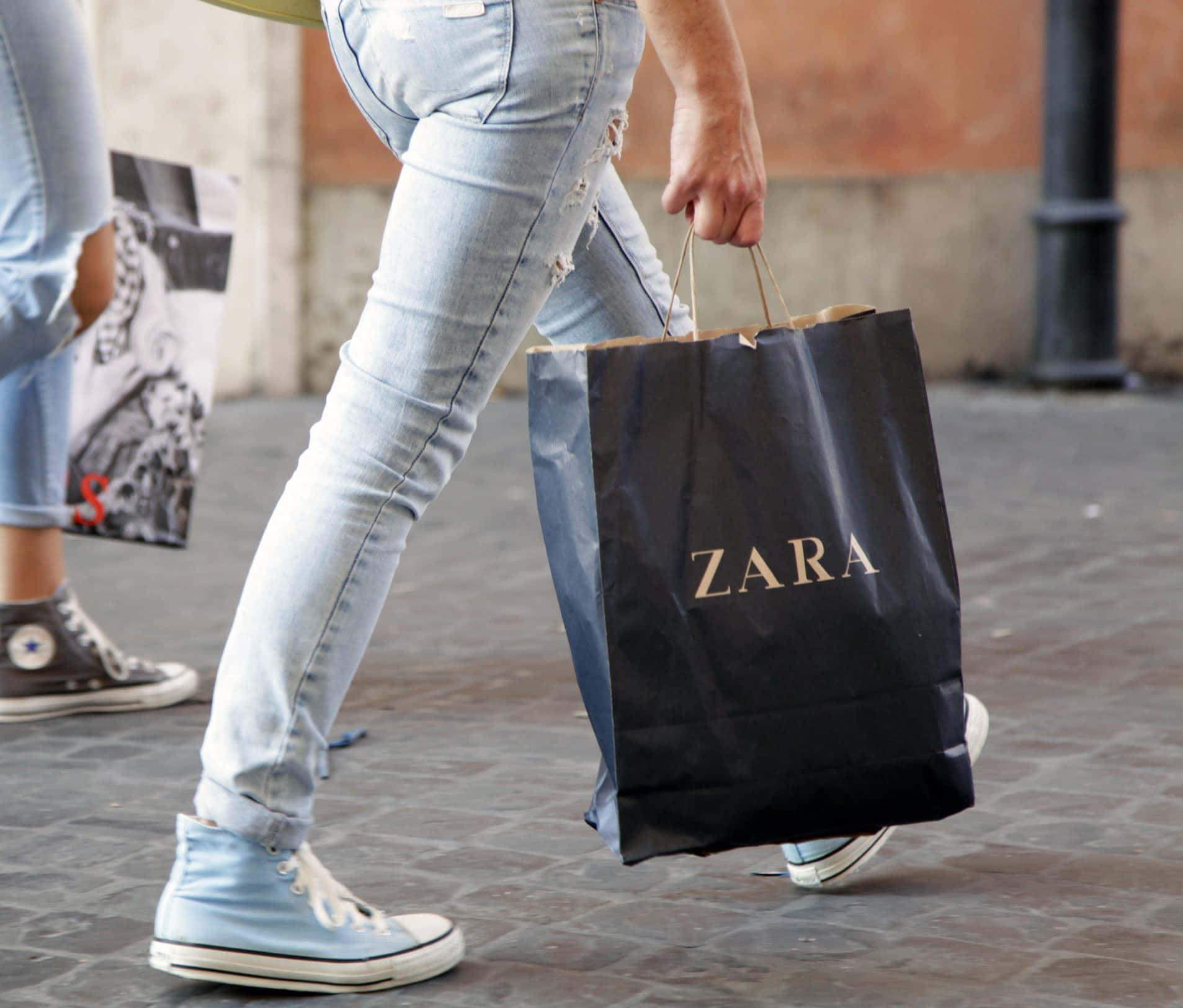 Zara最新系列 壁紙 — 一個穿著破洞牛仔褲和淺藍色運動鞋的人，在鋪砌的城市人行道上提著一個黑色的Zara購物袋行走。