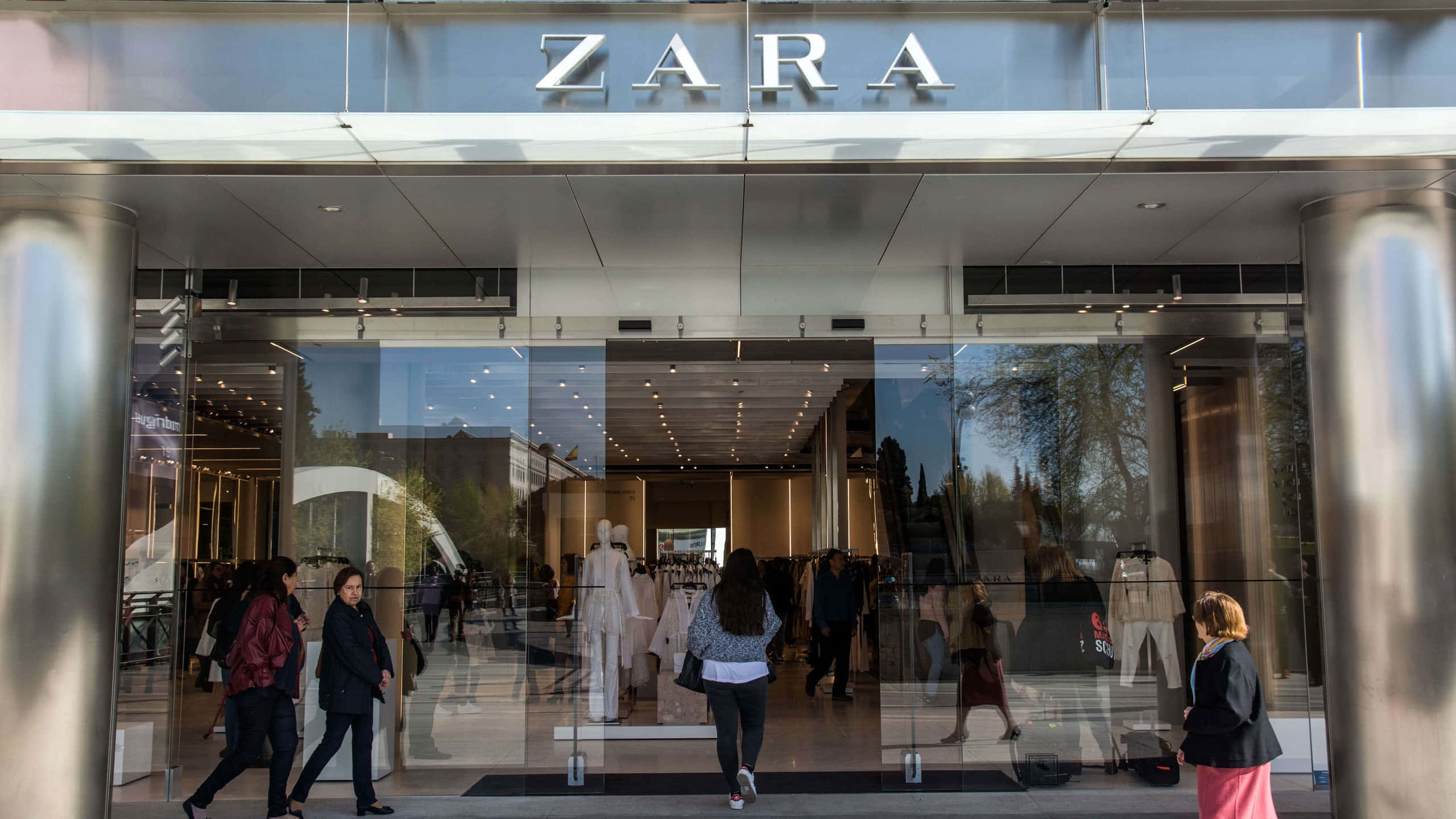 尋找適合冬季的Zara時尚造型 壁紙 — 人們走過一間現代化的Zara商店，玻璃門反射出樹木和店內瀏覽衣架的購物者。