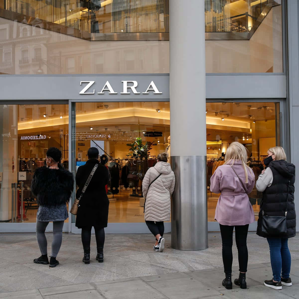 以Zara保持時尚 壁紙 — 人們在 Zara 商店外排隊，玻璃窗反射城市建築物和溫暖的室內燈光。