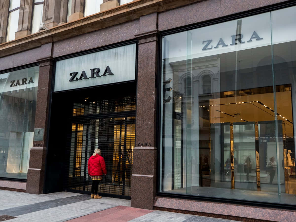 Zara：為所有人打造時尚 壁紙 — 一名身穿鮮紅色外套的人站在Zara店舖的玻璃入口前，店舖有反光窗戶和石造外牆。