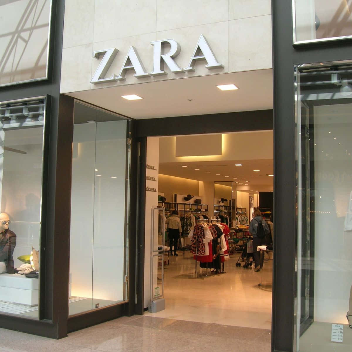 以Zara職業服飾展現時尚與專業 壁紙 — 現代Zara店舖入口，設有玻璃門和明亮的室內燈光，展示內部的衣架和購物者。