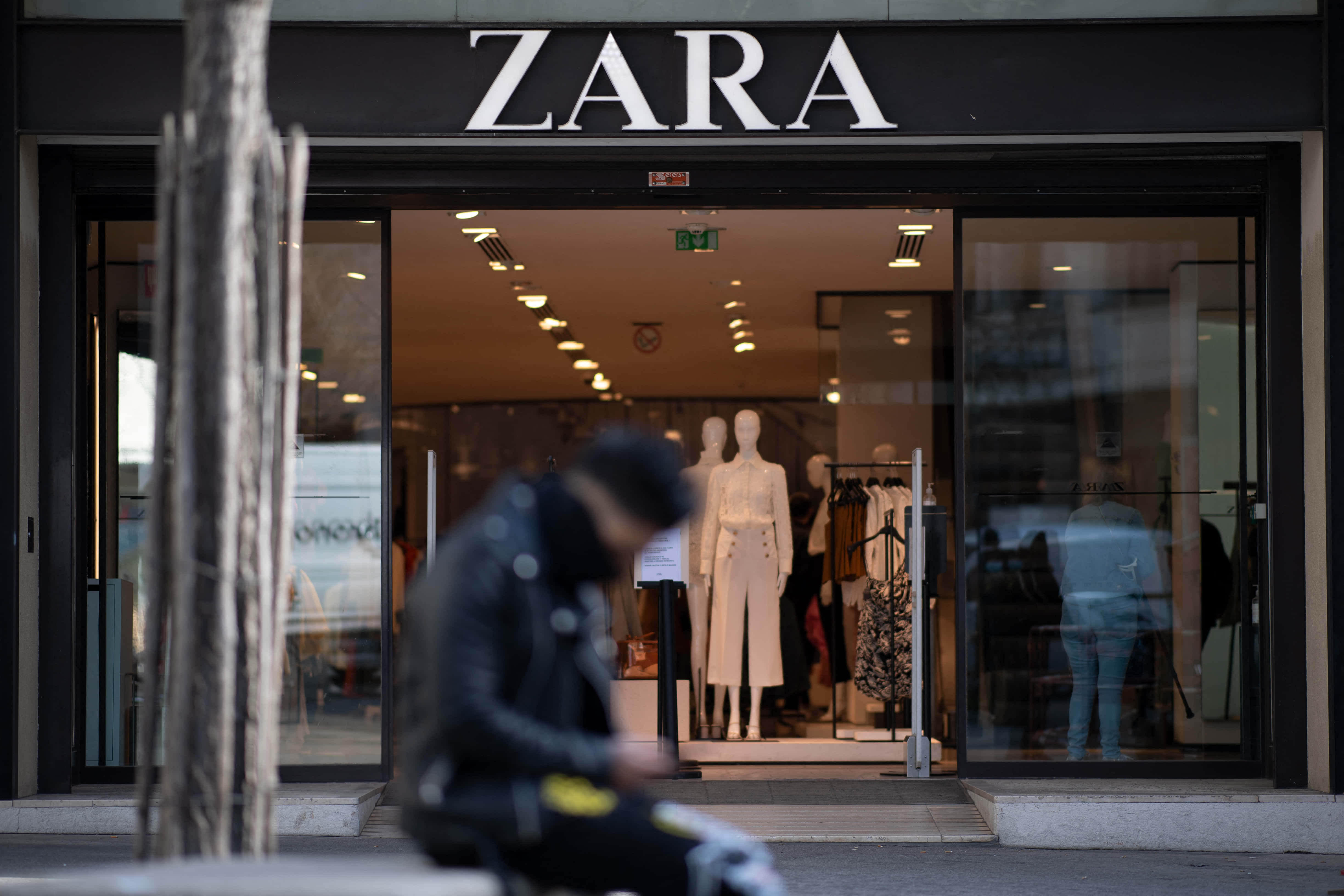 以Zara刷新衣櫥 壁紙 — 一個模糊嘅人著住黑色外套坐喺Zara舖頭外面，裏面有模特公仔展示白色服裝。