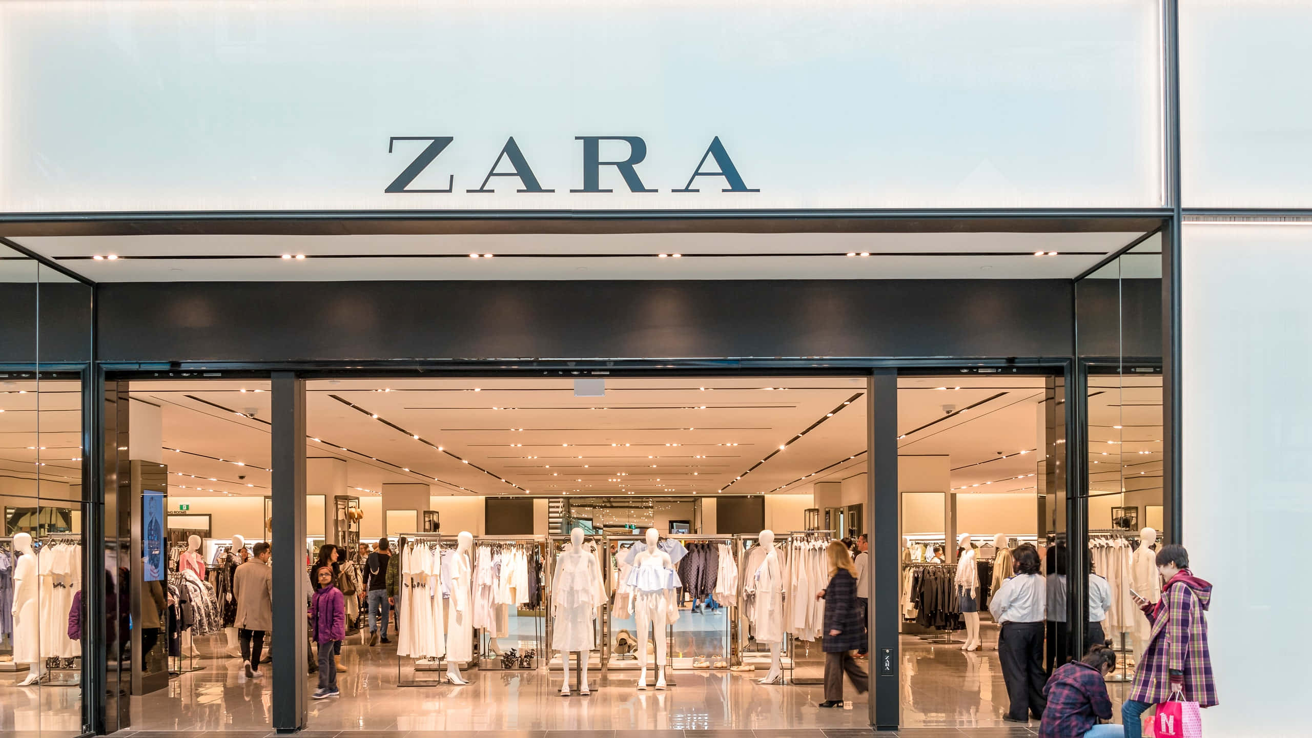 穿出風格，感受自信 — Zara 壁紙 — 明亮的Zara商店入口，玻璃門內可見人體模型和購物者瀏覽衣架。