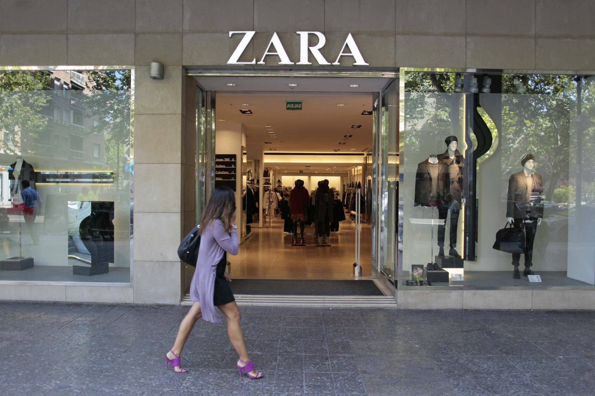 Zara時尚店外觀 壁紙 — 一位身穿紫色連衣裙和粉色高跟鞋的女性走過Zara商店，玻璃櫥窗內展示著人體模型和購物者。