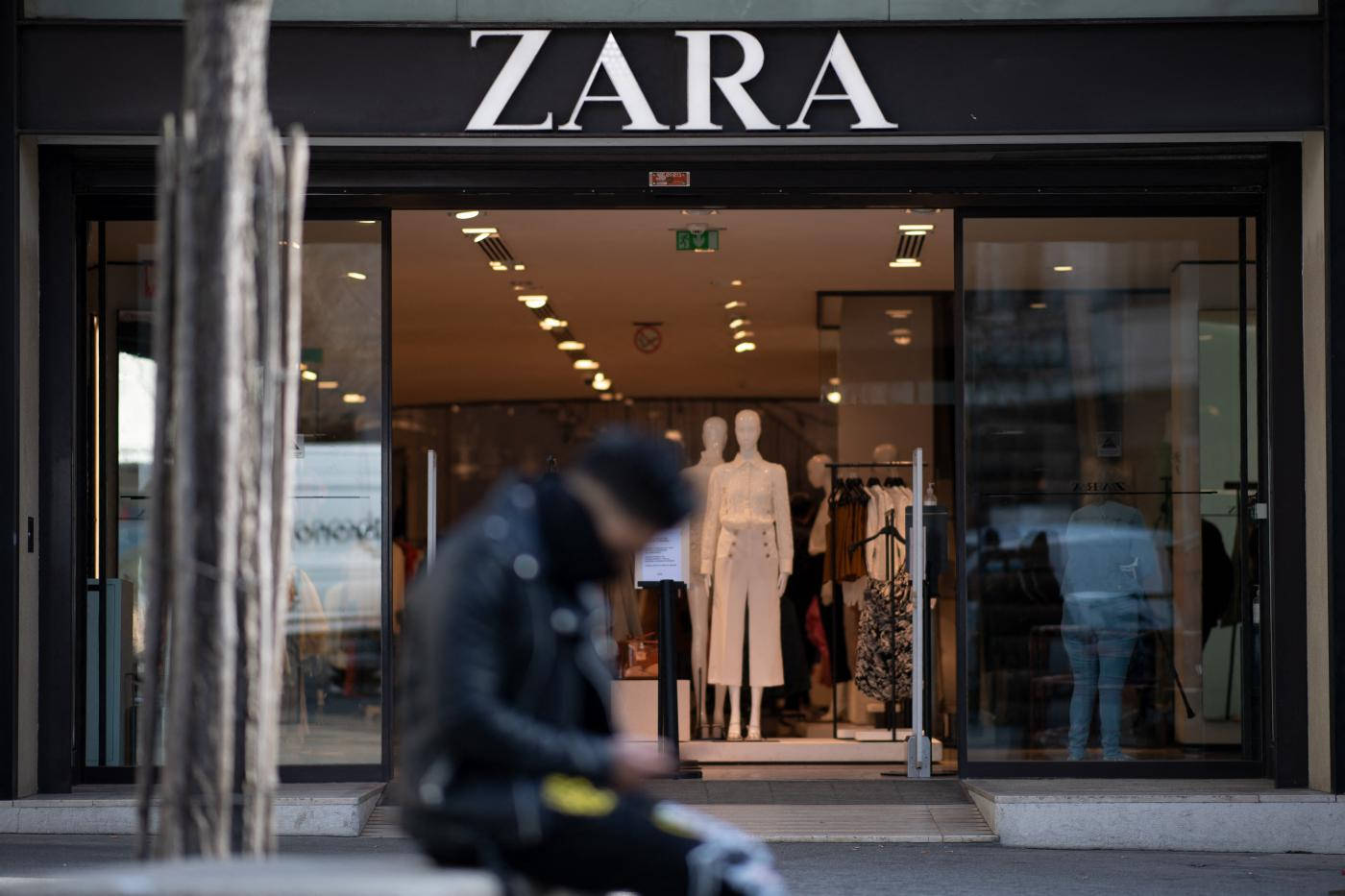 Zara服飾店門口 壁紙 — 一個模糊的人穿著黑色外套坐在Zara商店外面，玻璃門內可見人體模型和衣服架。