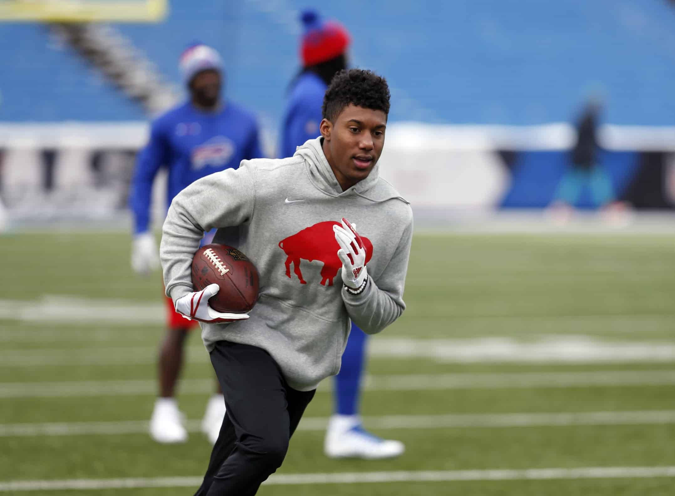 Zay Jones 灰色水牛連帽衫 壁紙 — 一位身穿灰色連帽衫、帶有紅色水牛標誌的年輕美式足球員，手持棕色美式足球在綠色球場上衝刺。