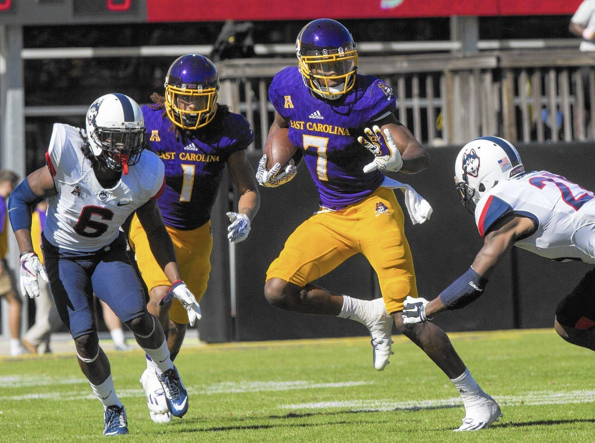 Zay Jones 動態比賽照 壁紙 — 一名身穿紫色和金色球衣的美式足球員持球奔跑，身穿白色和藍色球衣的對手在陽光明媚的球場上追趕他。