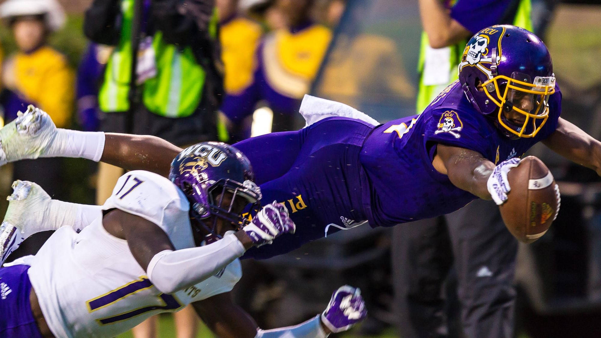 Zay Jones 撲球撞人 壁紙 — 一名身穿紫色球衣的足球員向前飛撲緊抱足球，同時一名白色球衣的防守員在半空中攔截他。