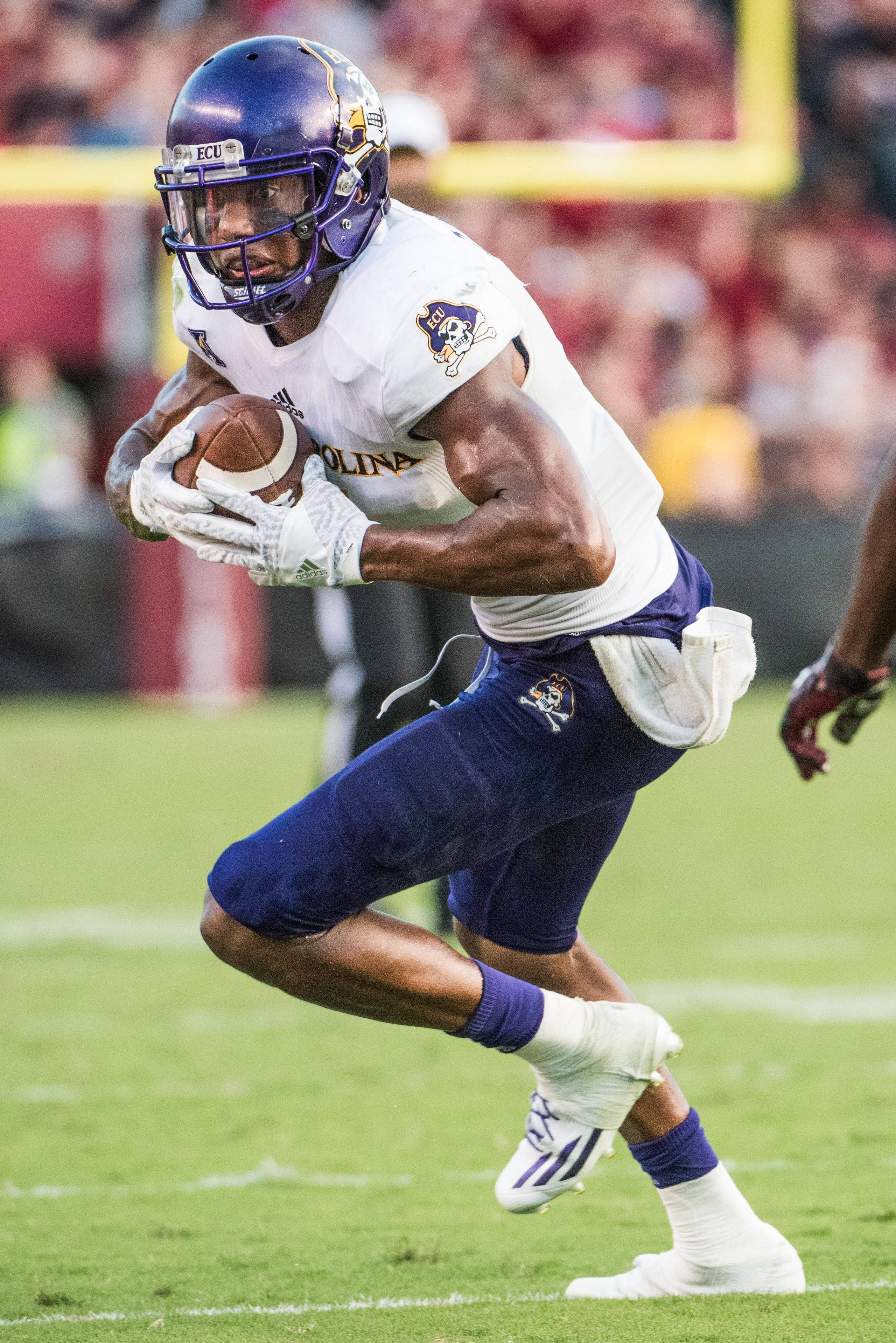 Zay Jones 球緊貼胸前 壁紙 — 一名身穿白色和紫色裝備的美式足球運動員在體育場燈光下的綠色球場上帶球衝刺。