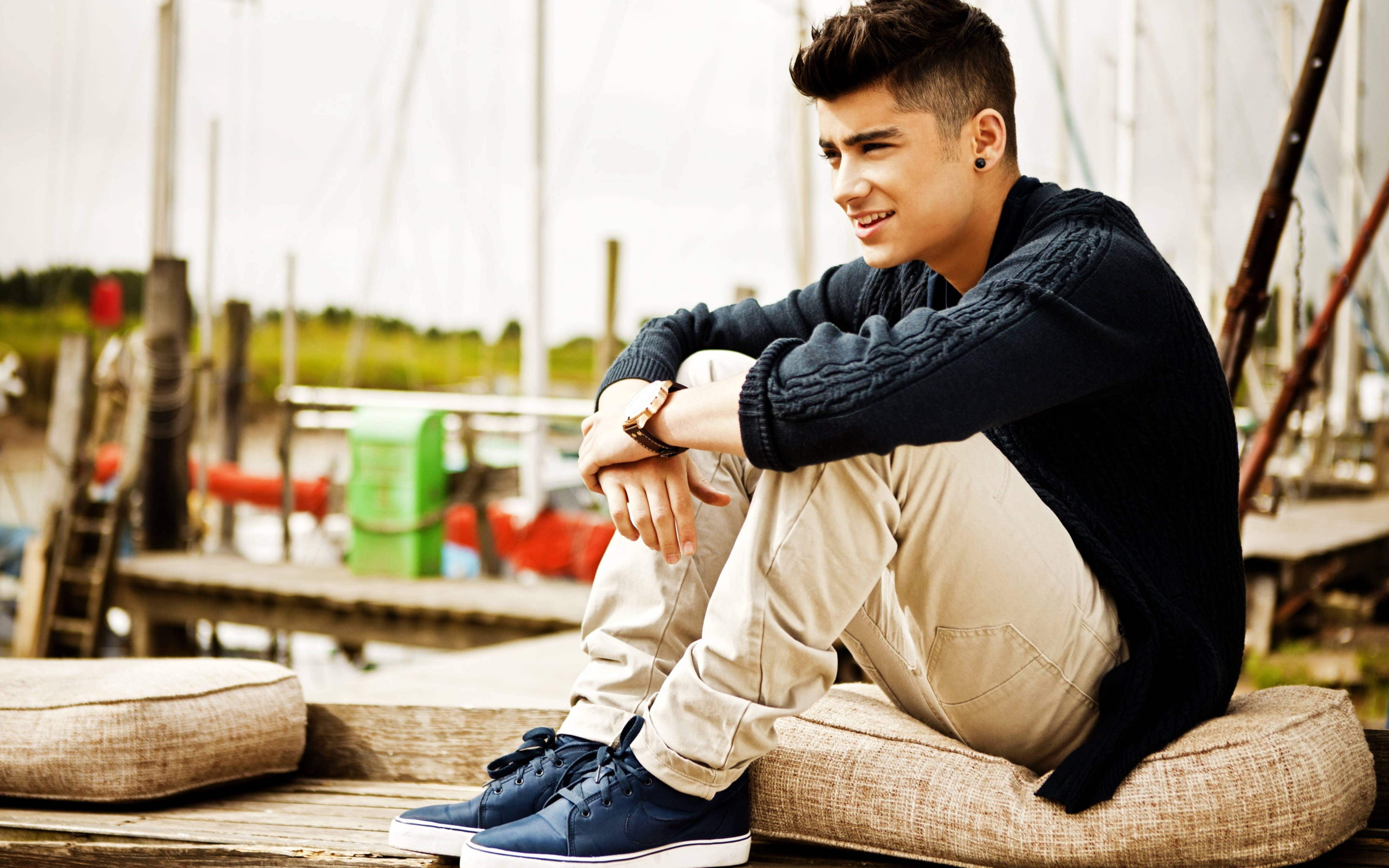 Zayn Malik 1D 拍攝時光 壁紙 — 一位年輕男子梳著時尚髮型，穿著深色毛衣和藍色運動鞋，隨意坐在碼頭坐墊上微笑。