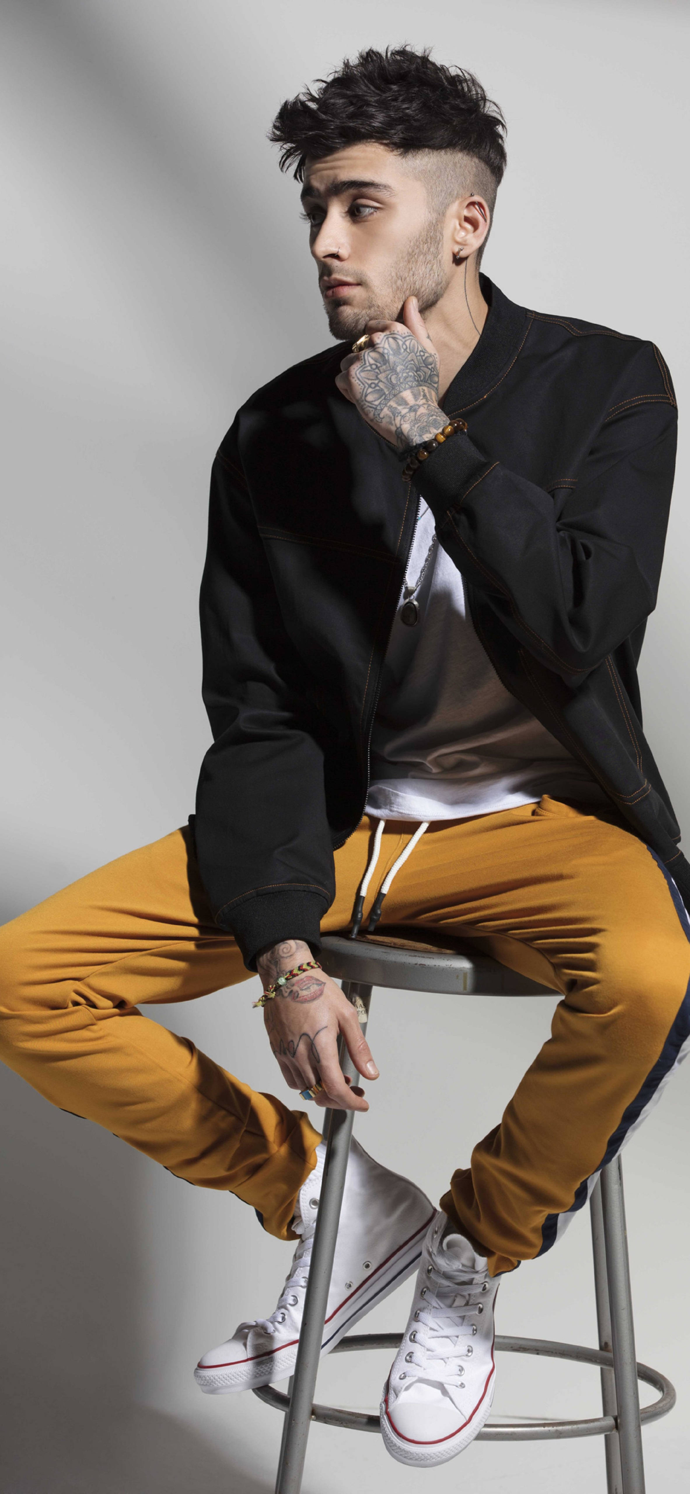 Zayn Malik Penshoppe Denimlab 2018系列 壁紙 — 一位時尚的年輕紋身男子坐在凳子上，身穿黑色外套、芥末色褲子和白色運動鞋，背景為純灰色。