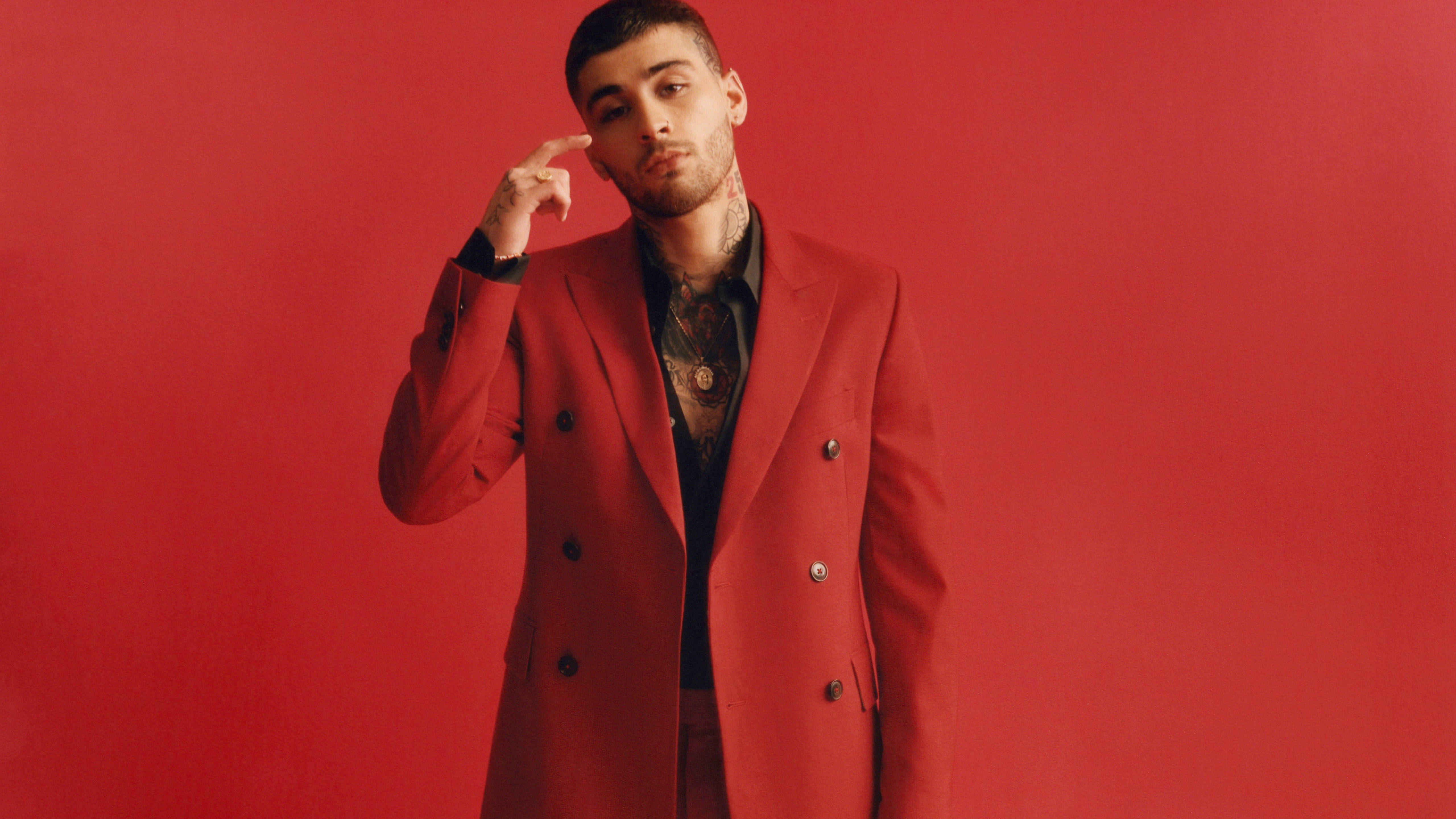 Zayn Malik 英國Vogue 壁紙 — 一名身穿大膽紅色雙排扣大衣的男子，手指抵著太陽穴，在純紅色背景前自信地擺姿勢。