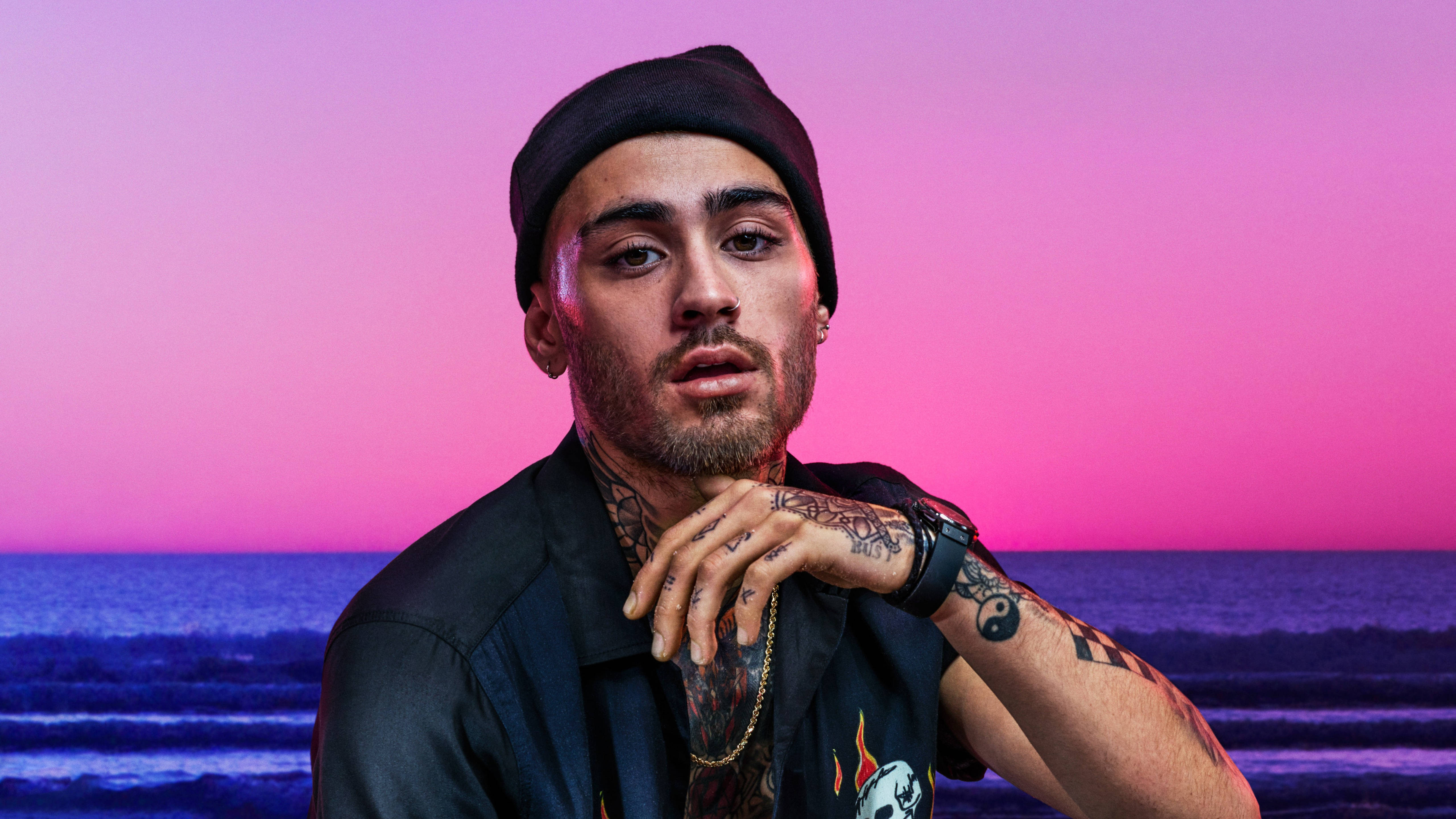 Zayn Malik為GQ雜誌2018年六月號拍攝的80年代復古風寫真 壁紙 — 一位紋身男子戴著黑色冷帽，在海洋上充滿活力的粉紅色和紫色日落前擺姿勢。