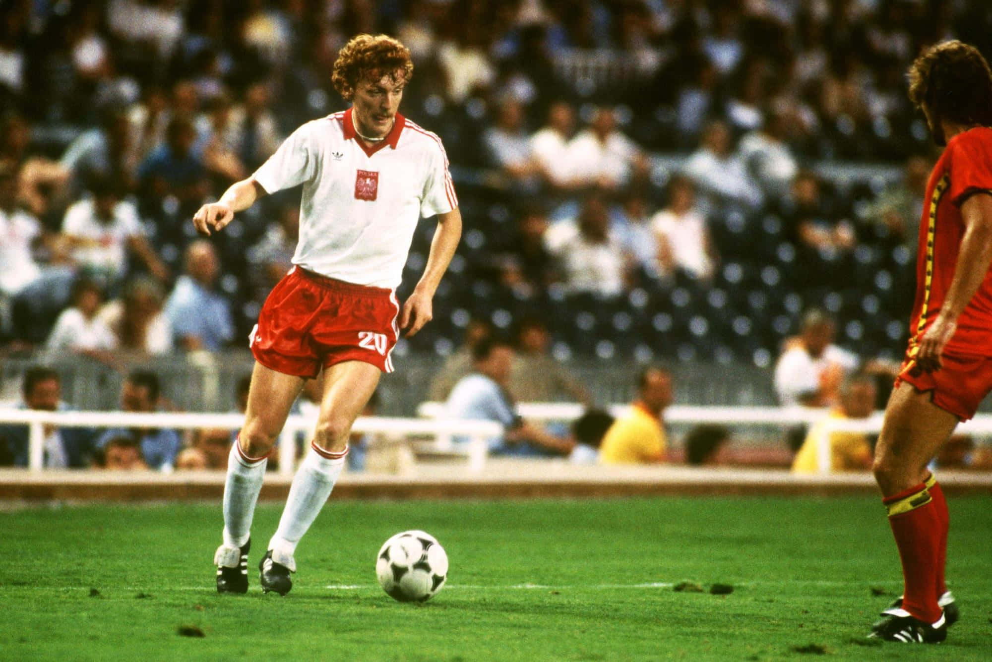 Zbigniew Boniek 在1982年世界杯波蘭對比利時賽事中控球 壁紙 — 一名身穿白色和紅色球衣的足球運動員在綠色球場上運球，背景有模糊的觀眾。