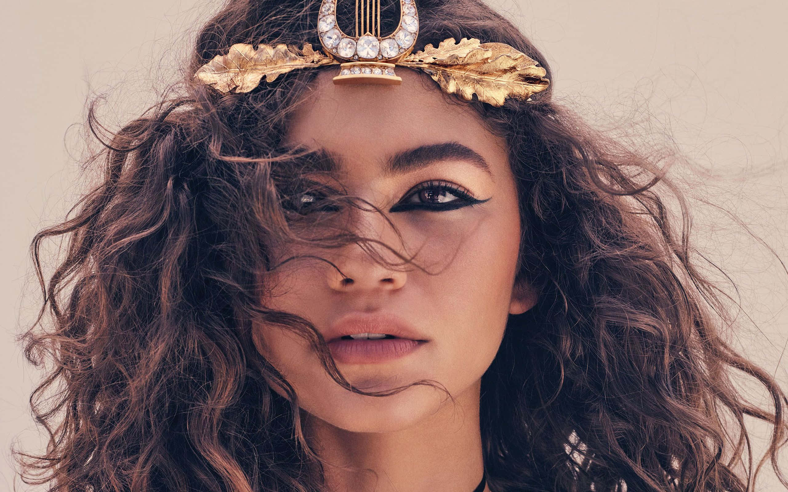 Zendaya 頭飾近距離高清照 壁紙 — 一位擁有棕色捲髮和戲劇性妝容的女性，在柔和的米色背景前佩戴金色月桂花環頭飾。