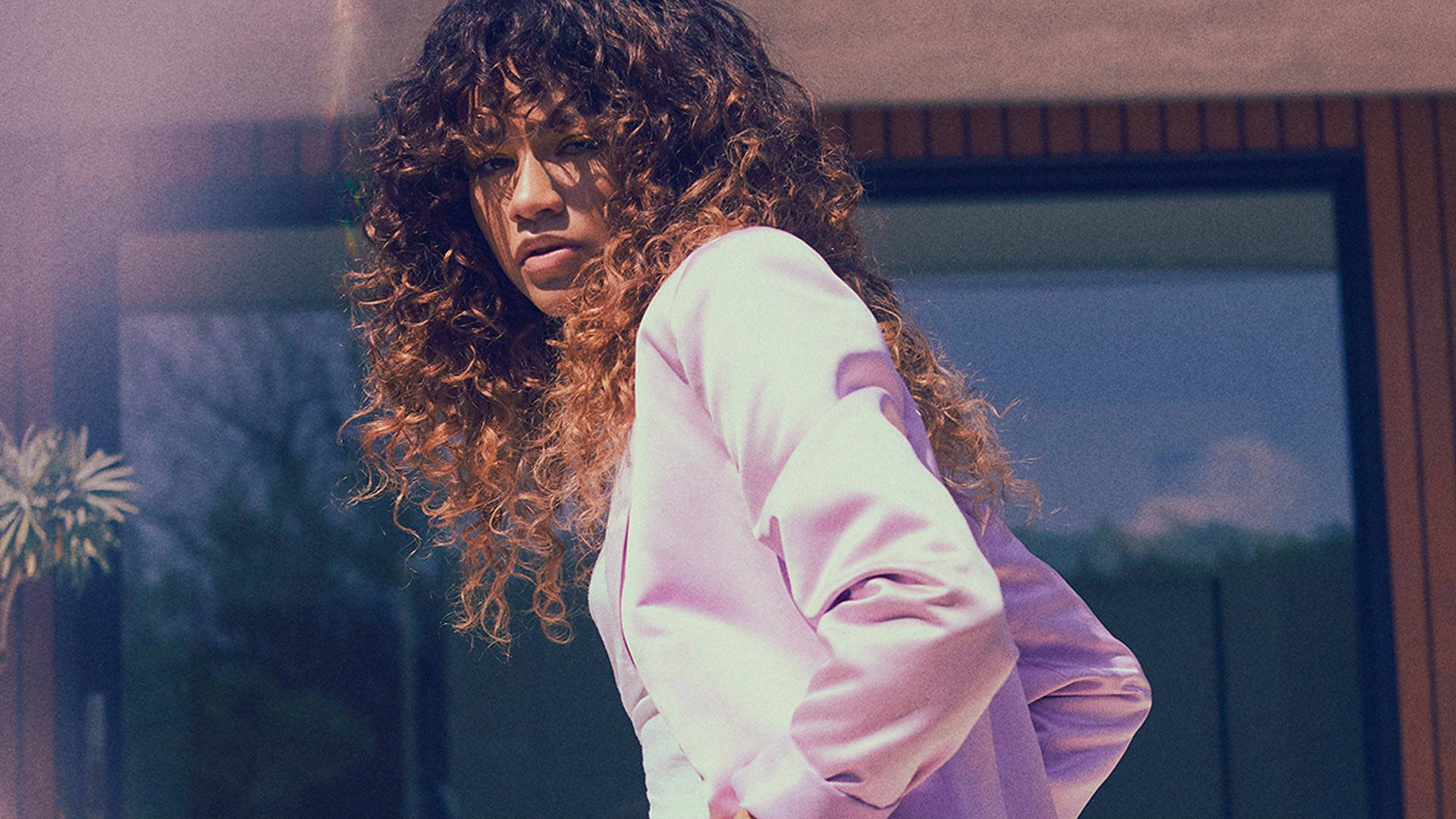 Zendaya x Boohoo 美學寫真 壁紙 — 一位擁有豐盈捲髮的女性穿著柔軟的粉紅色襯衫，站在現代玻璃建築附近的戶外。