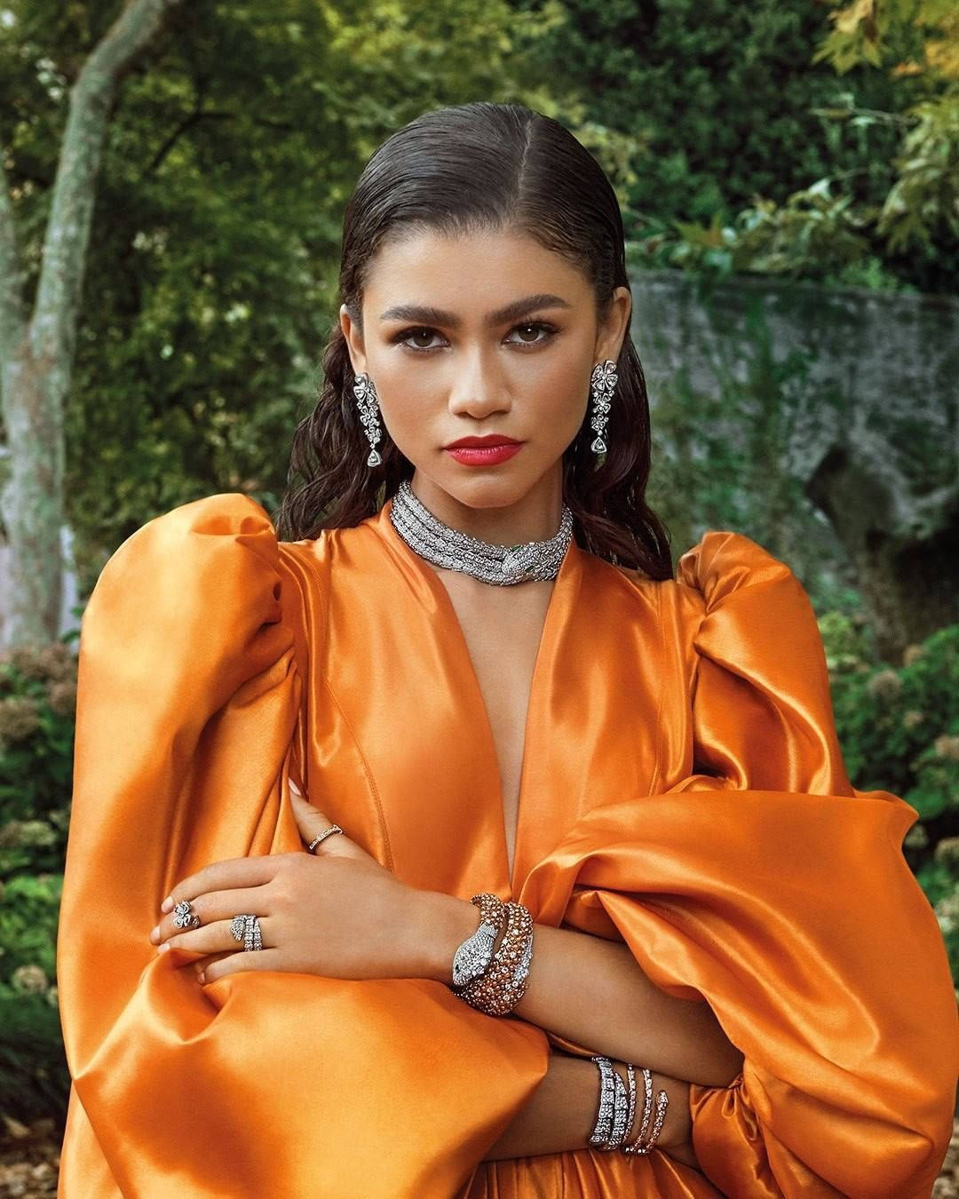 Zendaya x Bvlgari 珠寶寫真 壁紙 — 一位身穿鮮豔橙色緞面禮服、佩戴閃亮鑽石珠寶的女性，自信地站在茂密的綠色森林背景下。