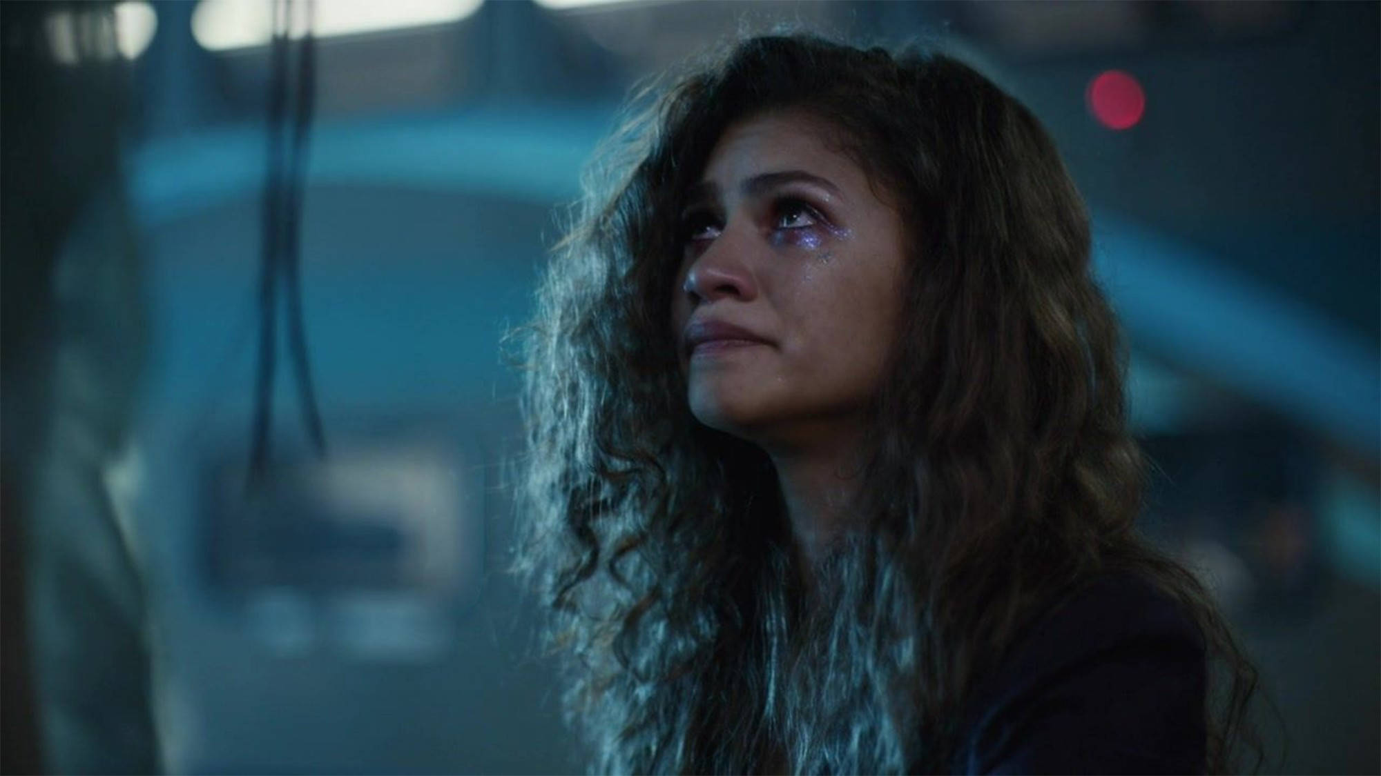 Zendaya 在《Euphoria》劇照 壁紙 — 一位捲髮女性在昏暗的未來場景中，在冷藍色燈光下哭泣。