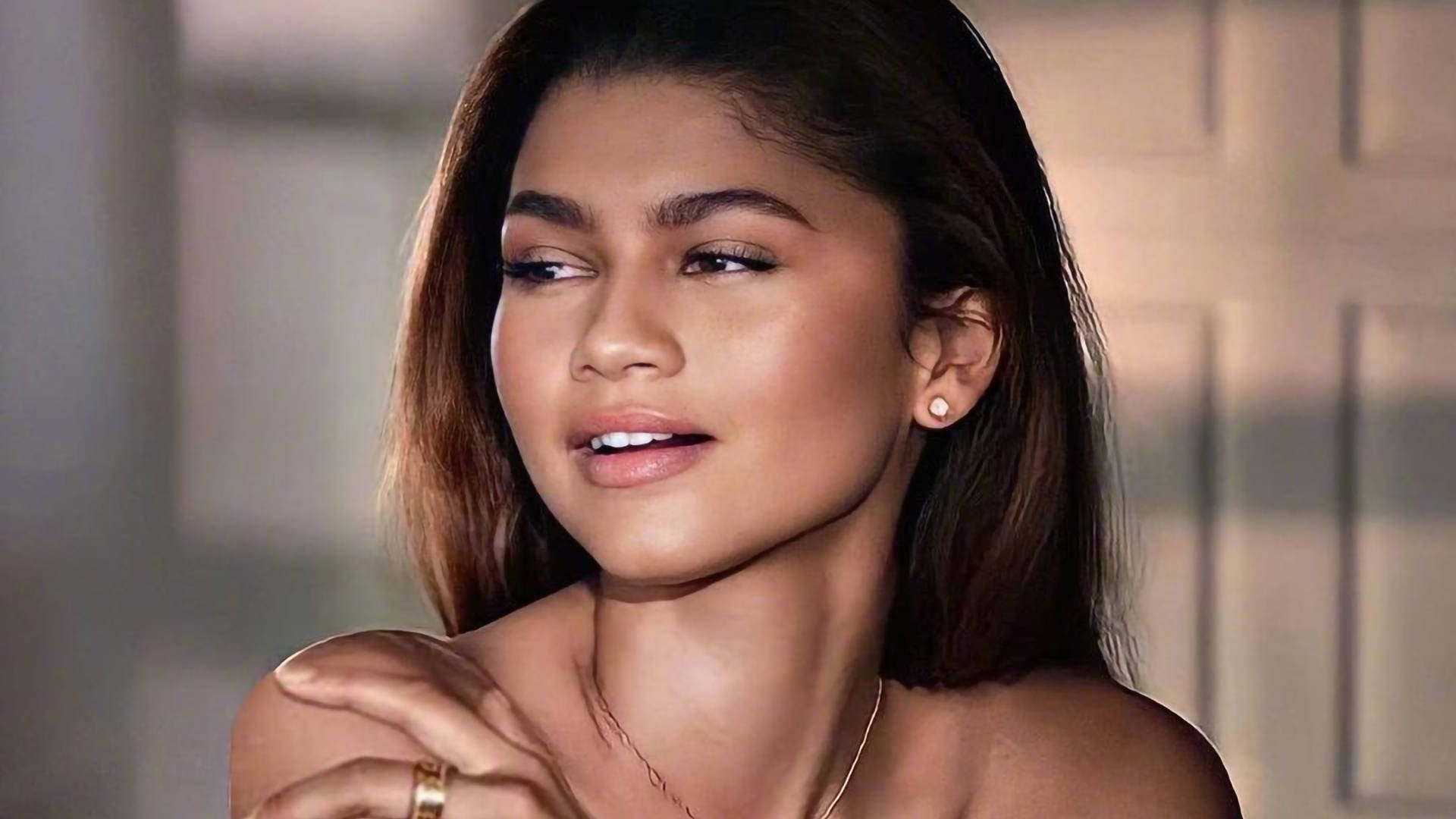 桌布 壁紙 — Zendaya 溫柔地凝視，帶著輕柔的微笑，佩戴著細緻的珠寶，背景柔和模糊。